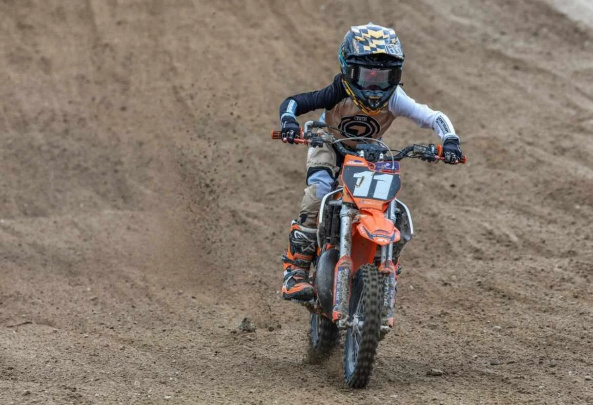 Young motocross rider wearing a helmet and protective gear riding an orange dirt bike on a dirt track.