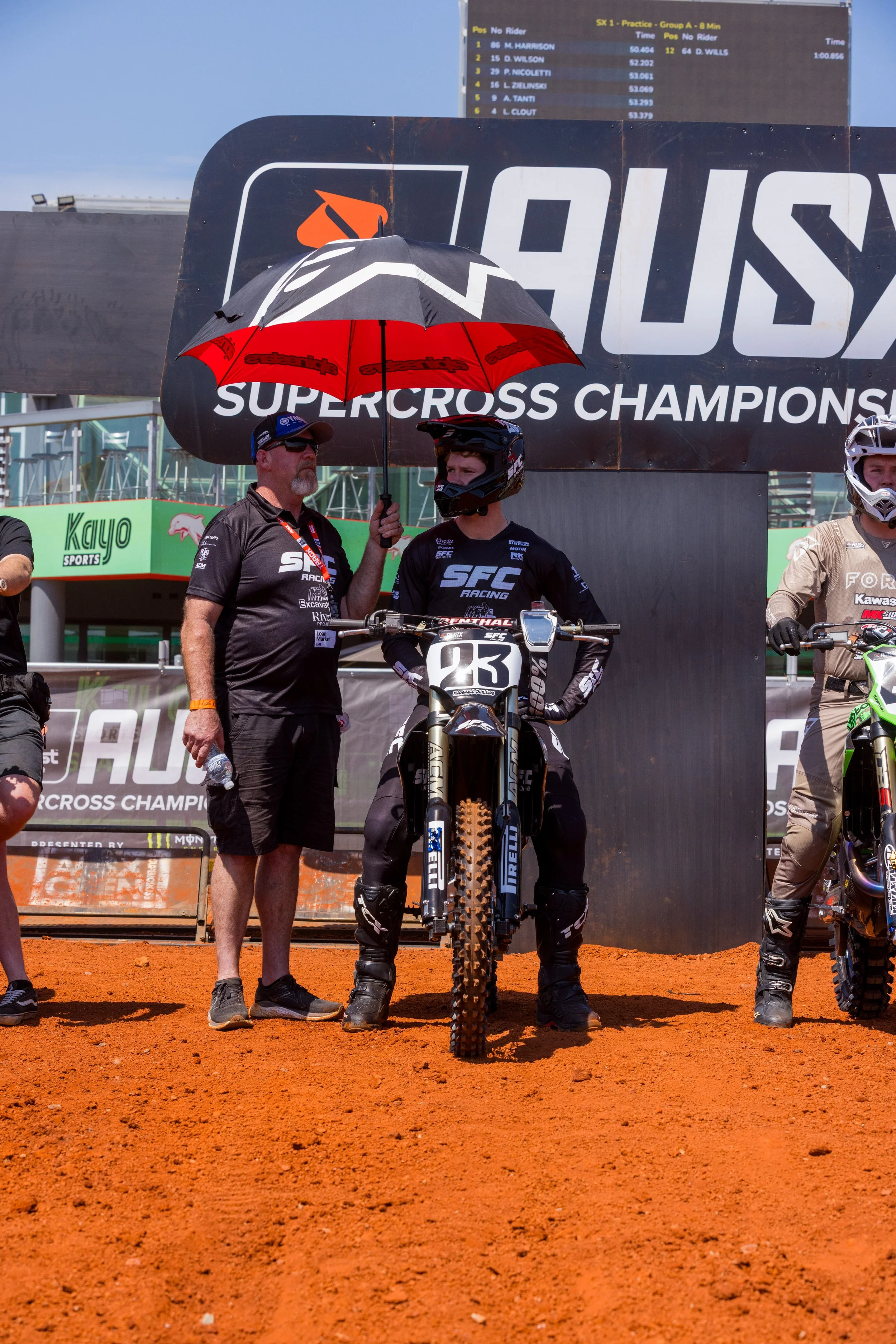 A motocross rider in black gear and helmet standing behind a dirt bike with race number 23, being talked to by a man holding an umbrella during a supercross event. The backdrop displays a large banner with the text 'Supercross Championships' and a leaderboard.