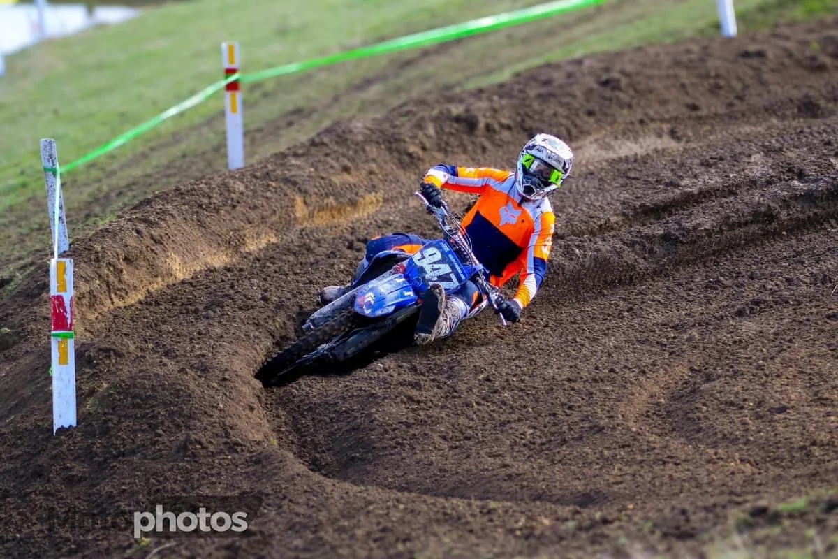 A motocross rider wearing an orange and blue jersey and helmet riding a blue dirt bike on a dirt track with a steep turn.