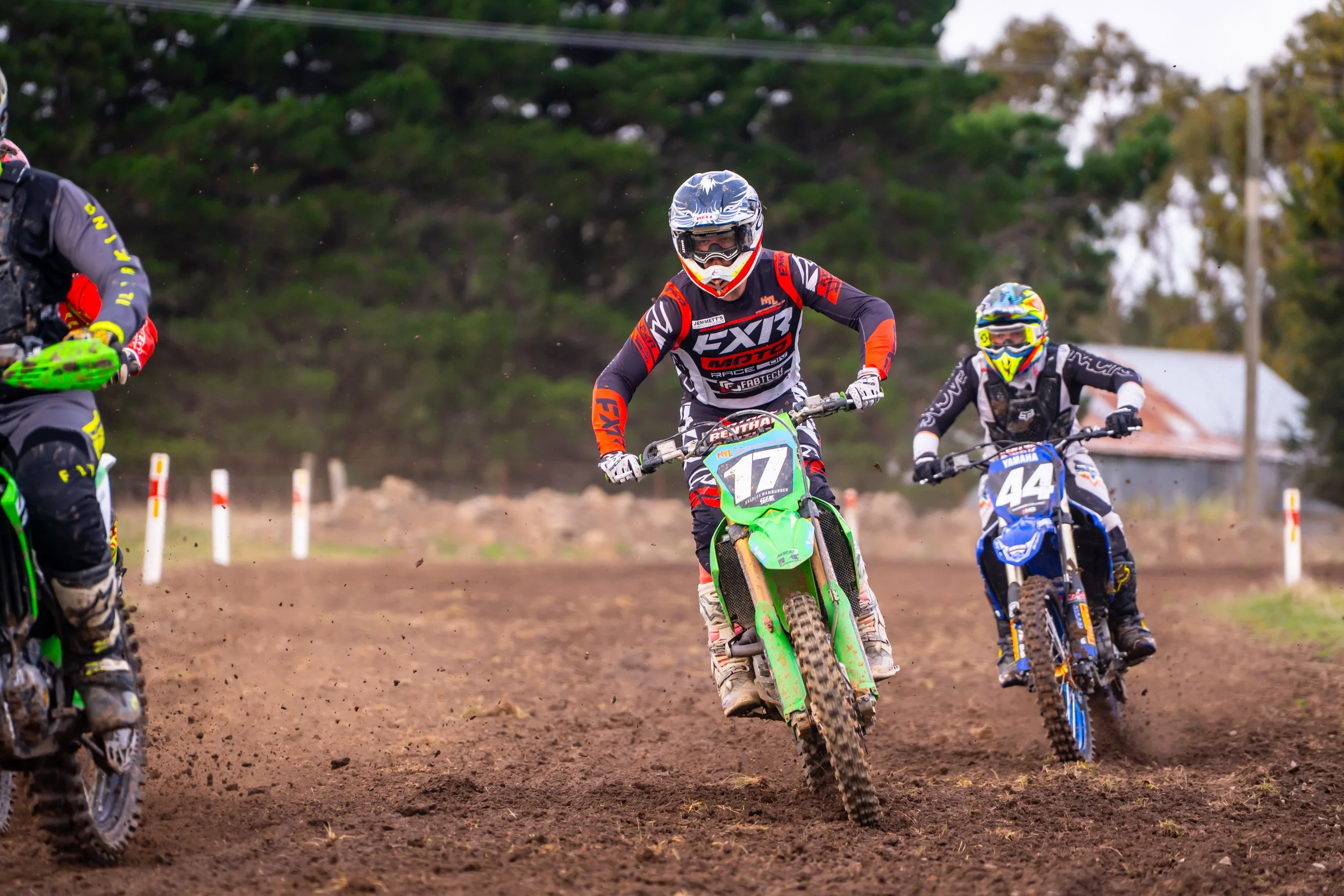 Motocross racers speeding on a dirt track, wearing colorful gear and helmets, with trees in the background.