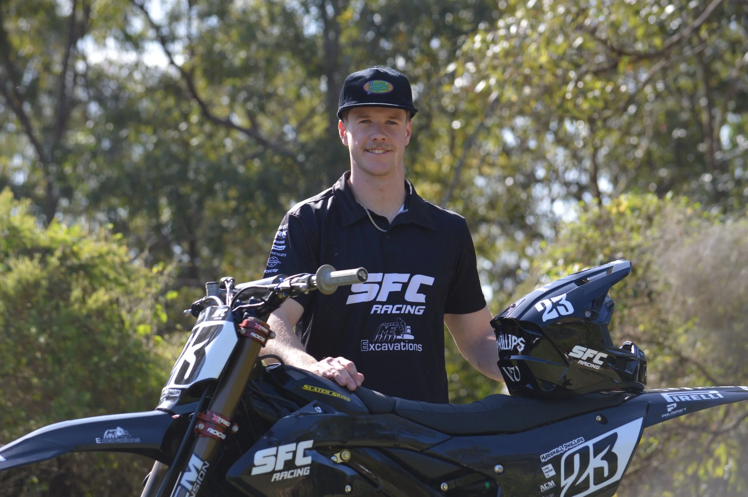 A young man wearing a black SFC Racing shirt and cap standing outdoors beside a black dirt bike with the number 023, holding a matching black helmet. The background shows trees and sunlight.