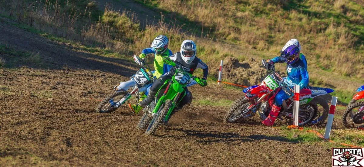Three motocross riders wearing colorful gear and helmets racing on a dirt track, kicking up dust as they lean into a turn.