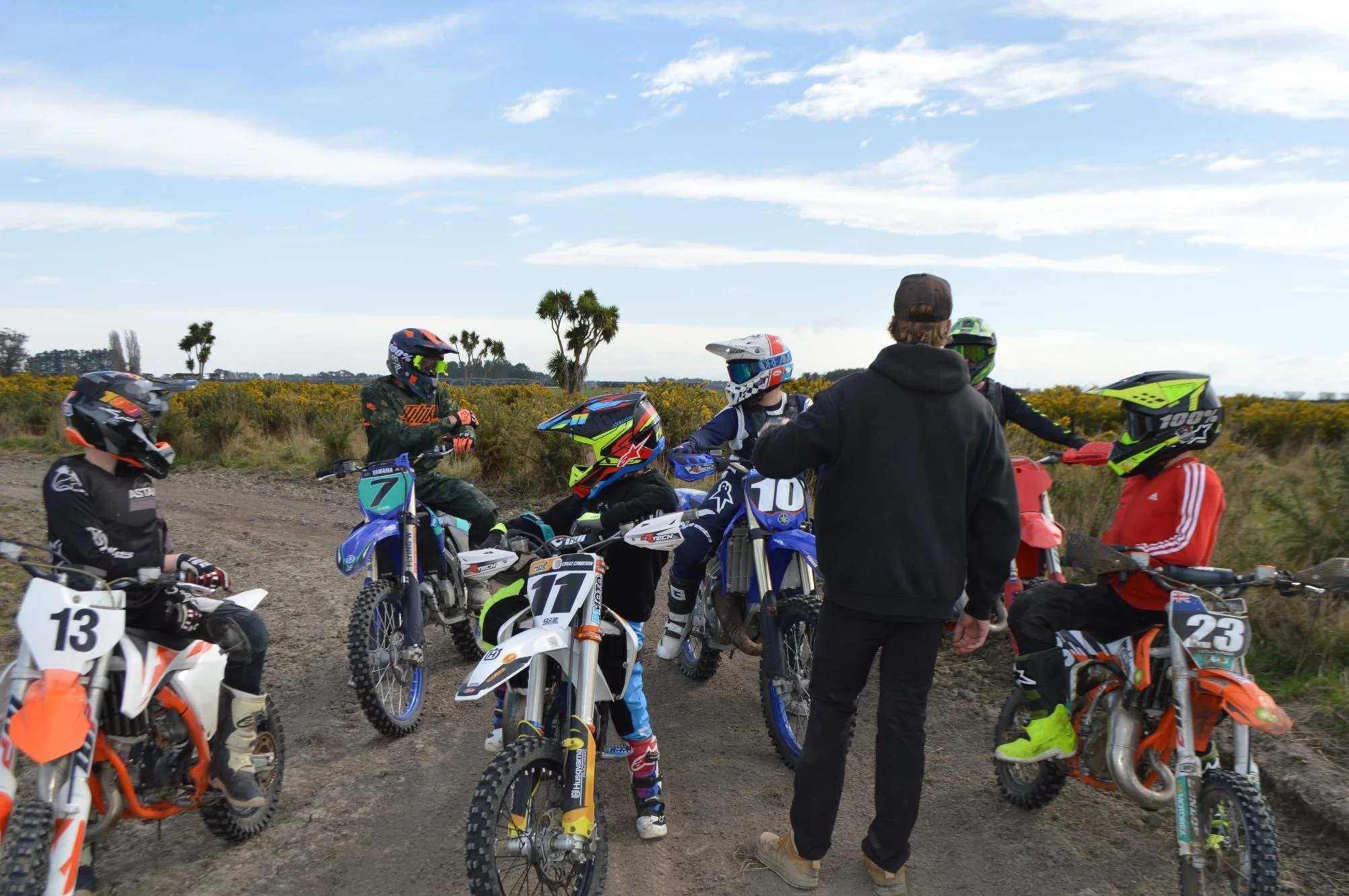 Group of dirt bike racers in riding gear and helmets gathered on a dirt trail, with one person standing and talking to the others, under partly cloudy skies with sparse vegetation and trees in the background.