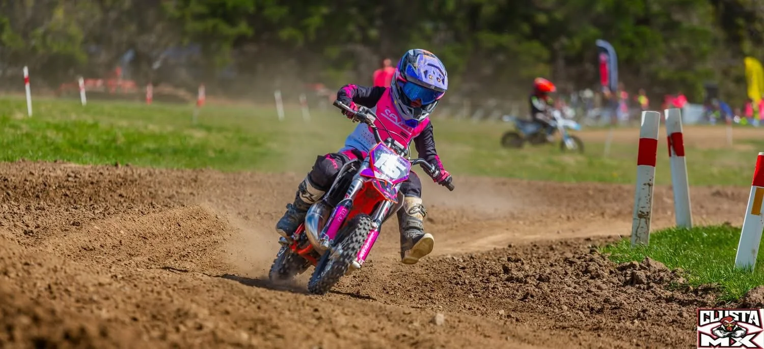 A young person riding a dirt bike on a race track, wearing a blue helmet and pink racing gear, leaning into a turn on a dirt track with grass and other riders visible in the background.