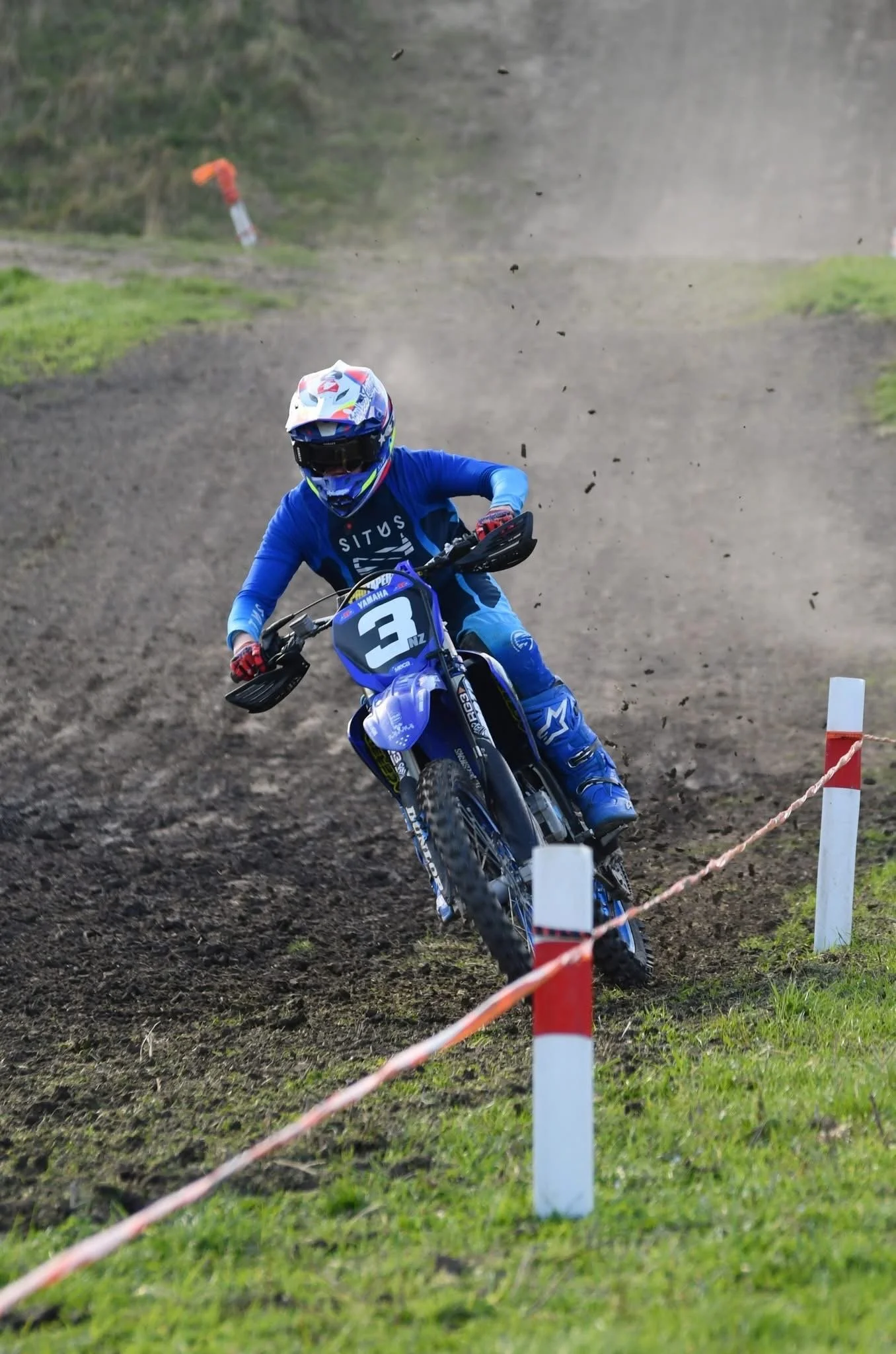 A motocross rider wearing a blue uniform and helmet riding a blue dirt bike on an off-road track, kicking up dirt.