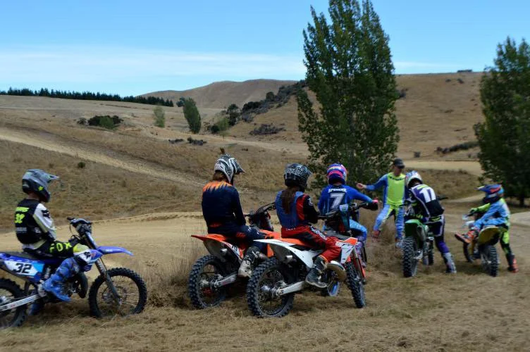 Group of children and an adult with helmets and motocross gear, with dirt bikes on a grassy field in a rural landscape with rolling hills and a few trees.