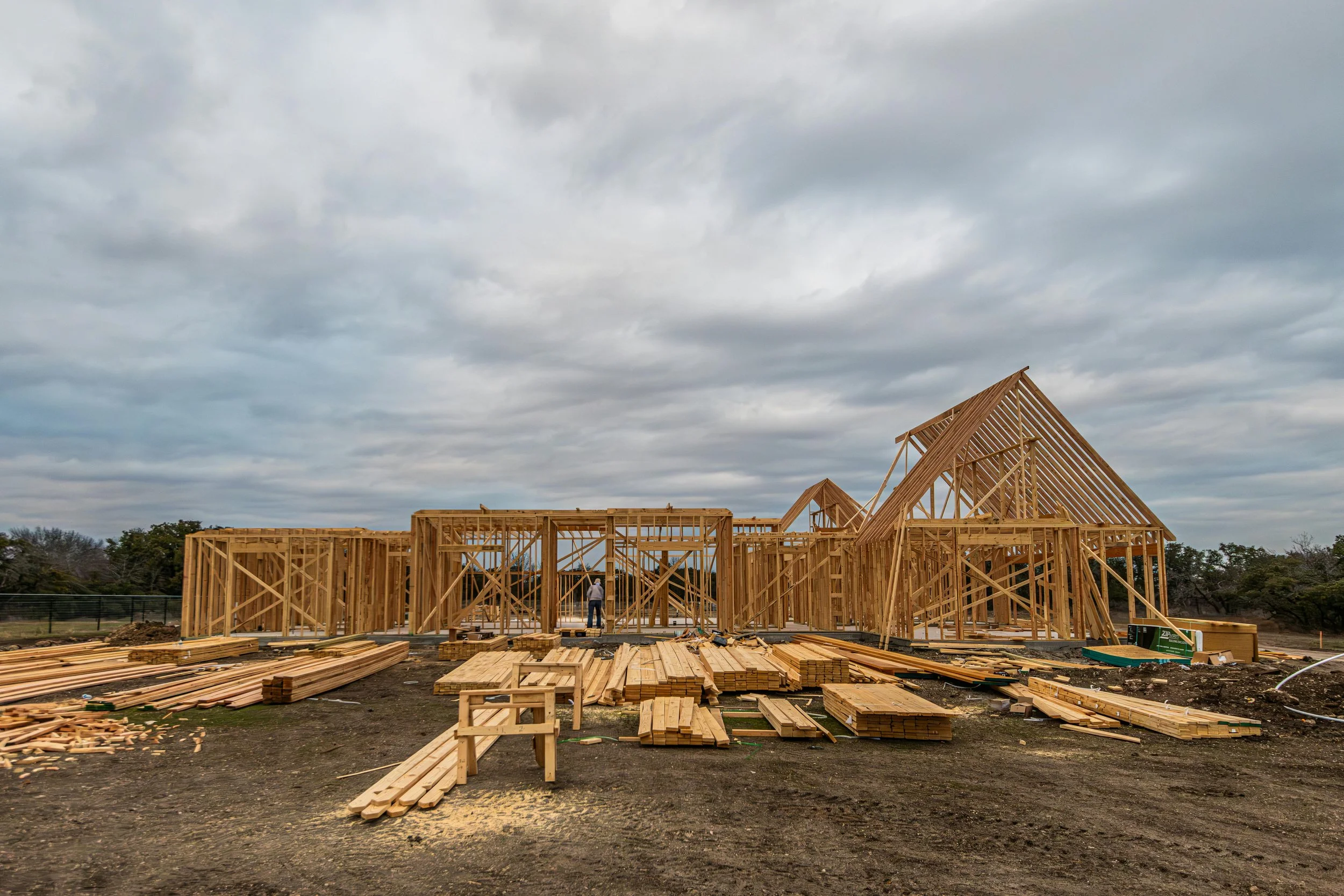 Construction site with wooden framing for a house, with building beams and lumber scattered on the ground, under a cloudy sky.