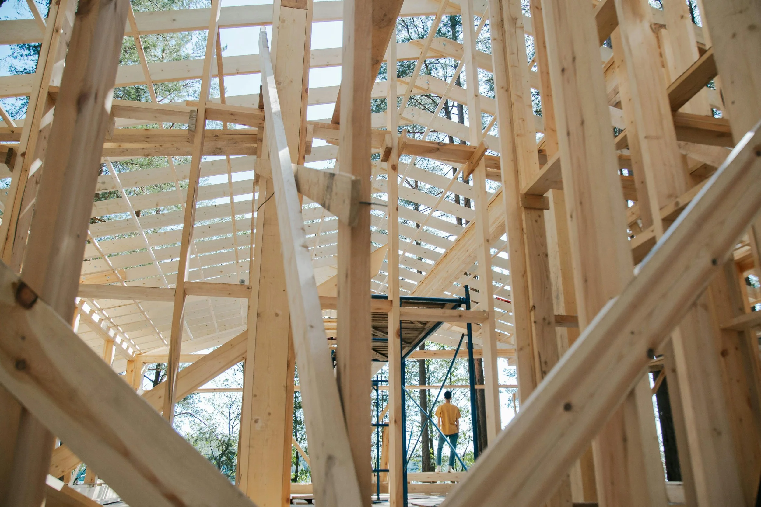 Wooden framing of a building under construction, scaffolding and a worker in a yellow shirt visible.