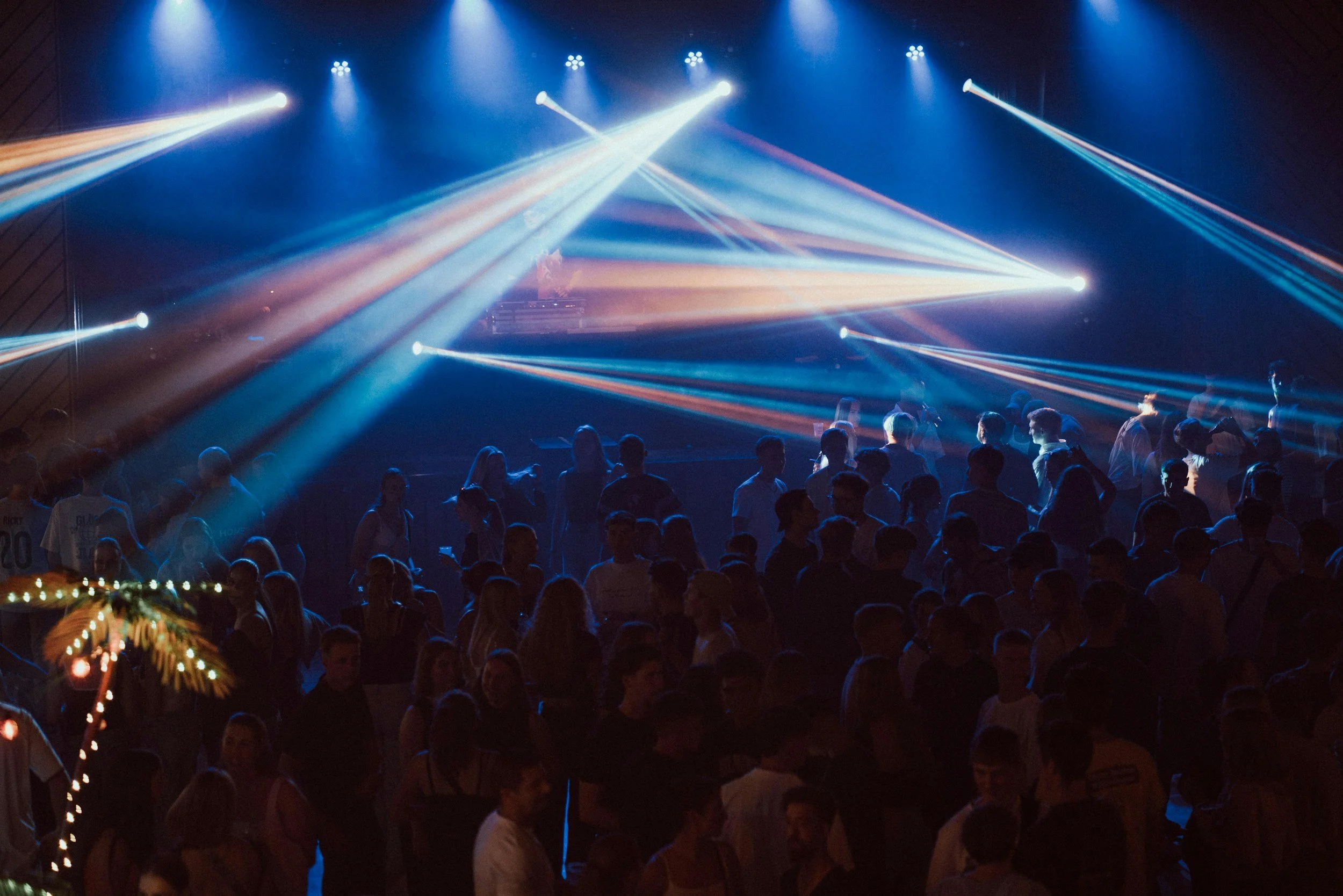 A crowded dance floor with people under colorful stage lights and moving beams of light in a dark venue.
