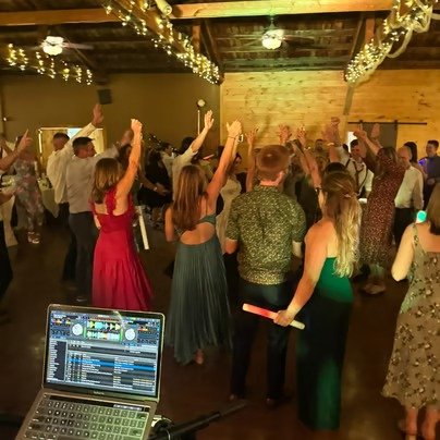People dancing and having fun at a party or celebration with a DJ setup and string lights hanging from the ceiling.