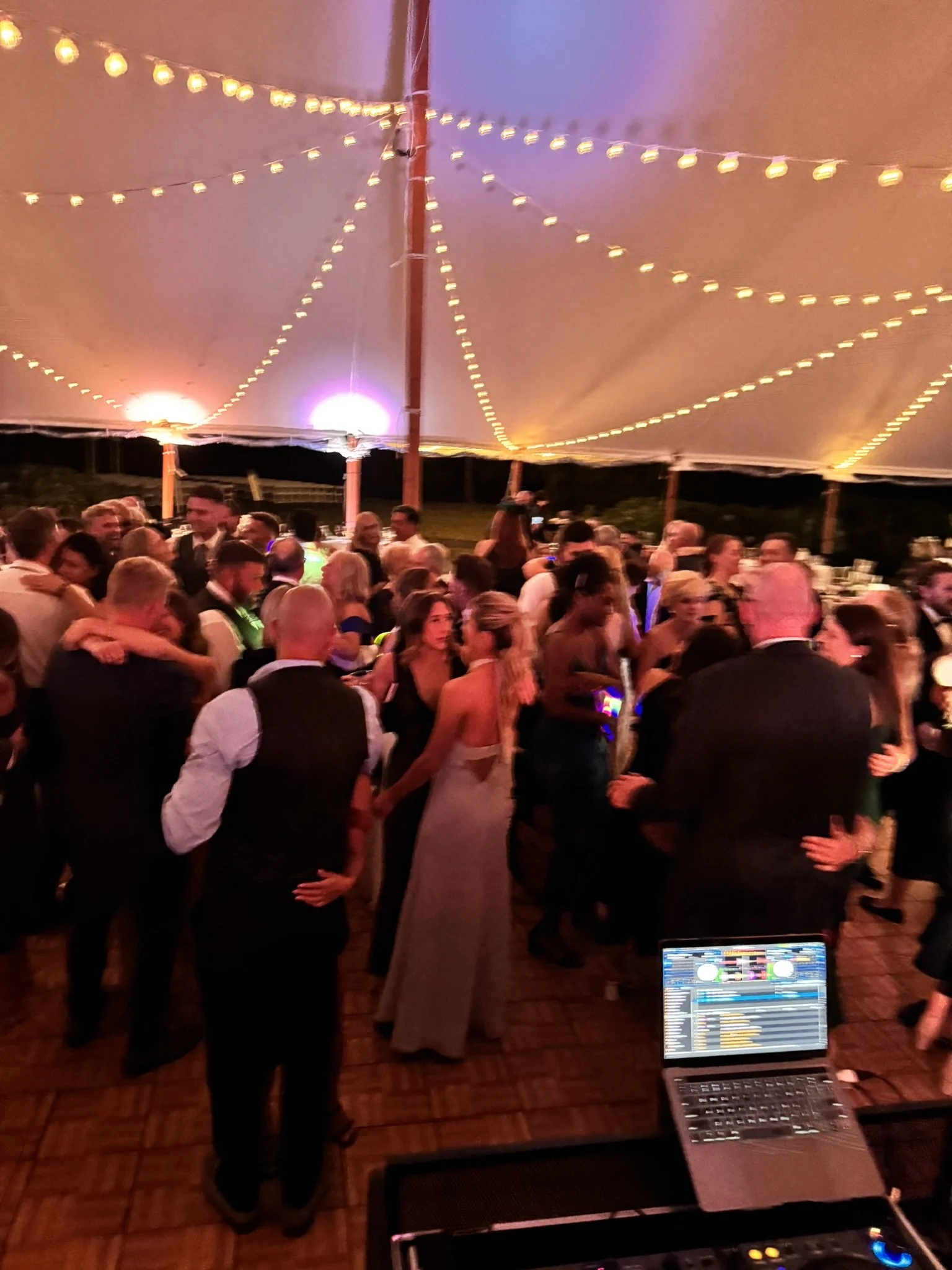 People dancing and socializing under a tent decorated with string lights at a wedding reception or party, with a DJ setup visible in the foreground.