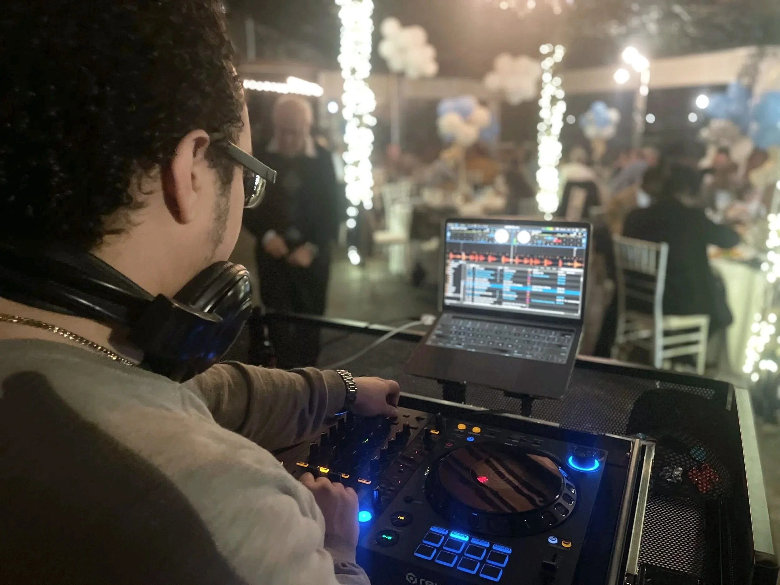 DJ at a wedding reception with a laptop and DJ equipment, with guests seated at decorated tables in the background.