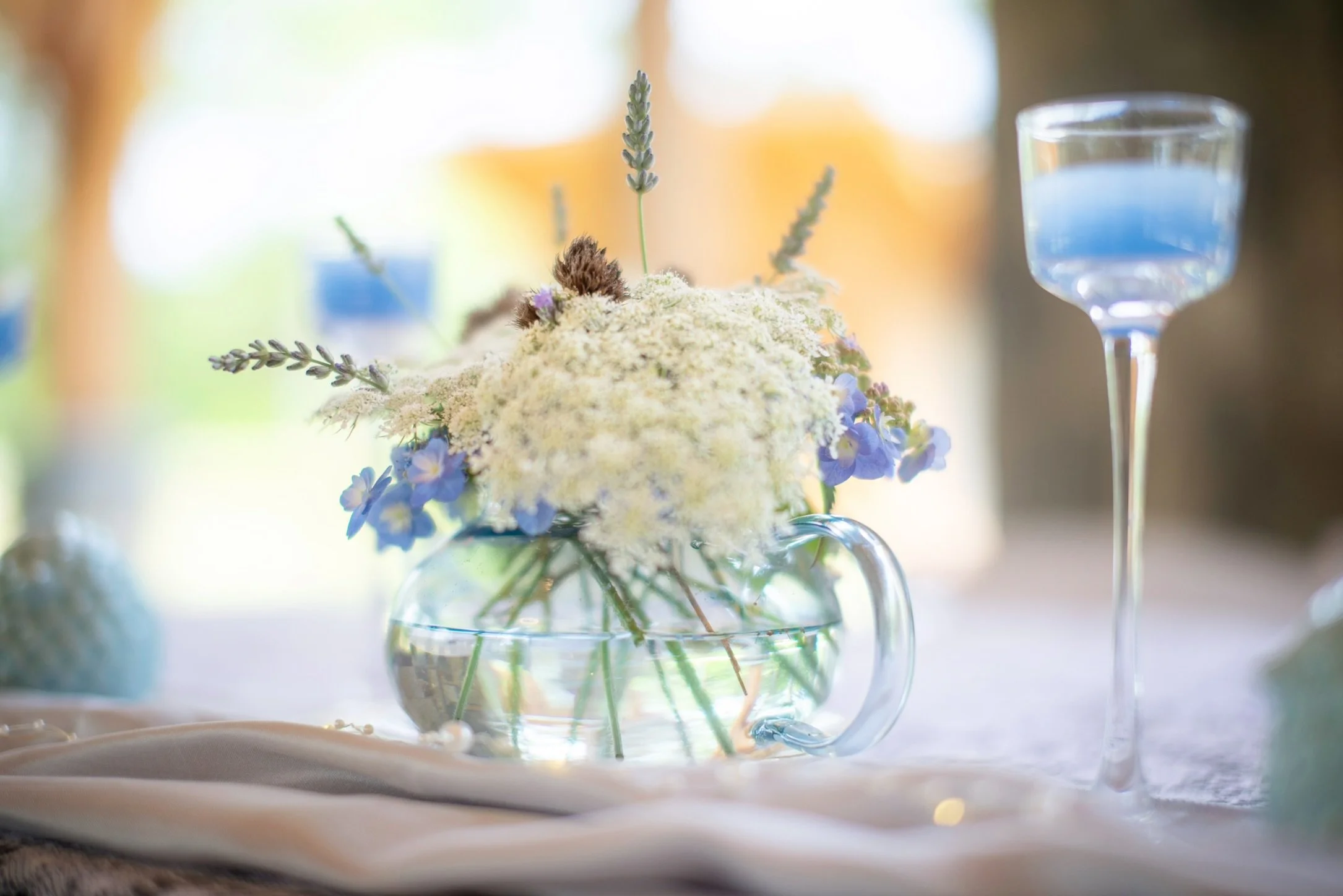 A floral centerpiece in a glass pitcher with white, purple, and lavender flowers, on a table with a white tablecloth, with a blue drink in a tall glass nearby, blurred background with warm lighting.