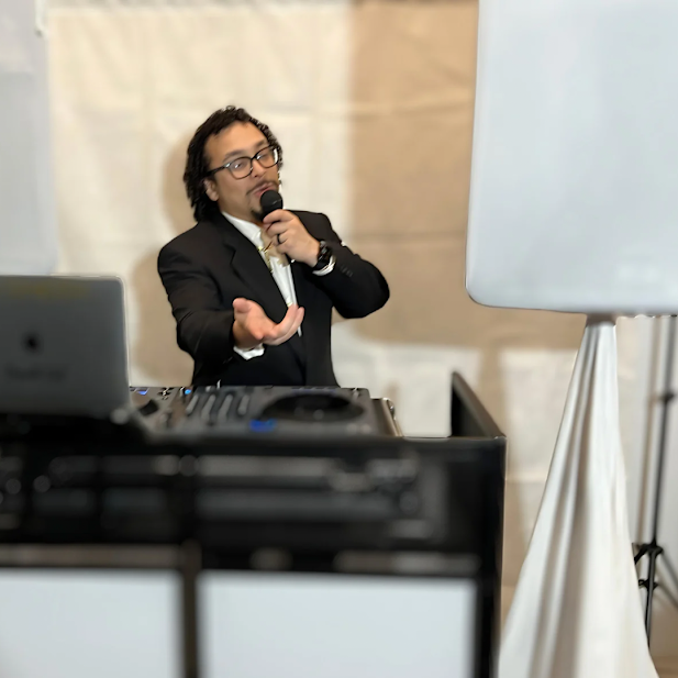 DJ Sneek talking on a microphone with his hand out pointing to the crowd behind his dj booth with speakers