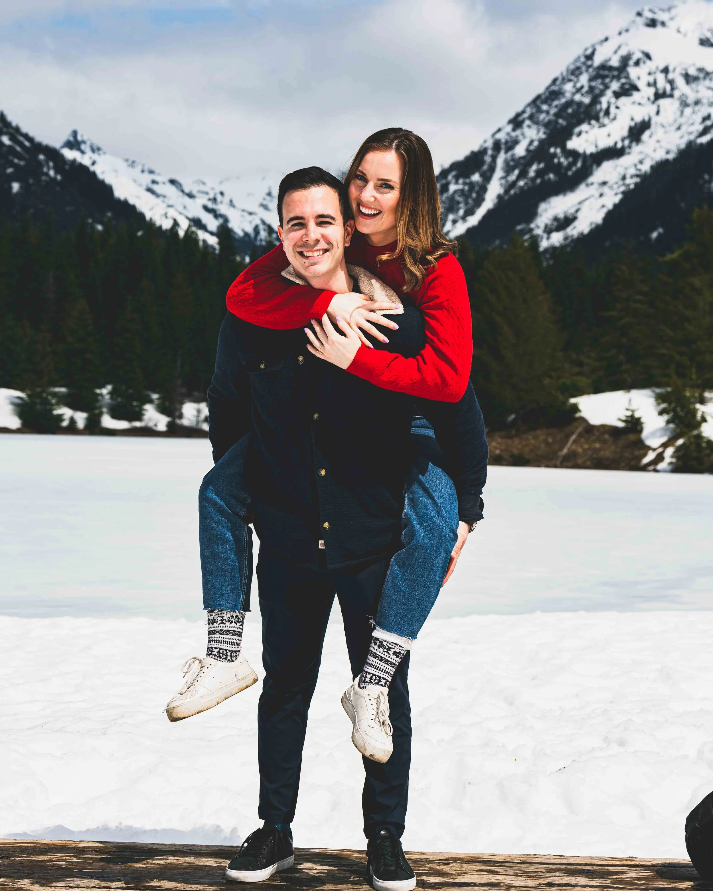A man giving a piggyback ride to a woman on a snowy mountain landscape with snow-covered peaks and a forest of evergreen trees in the background, both smiling.