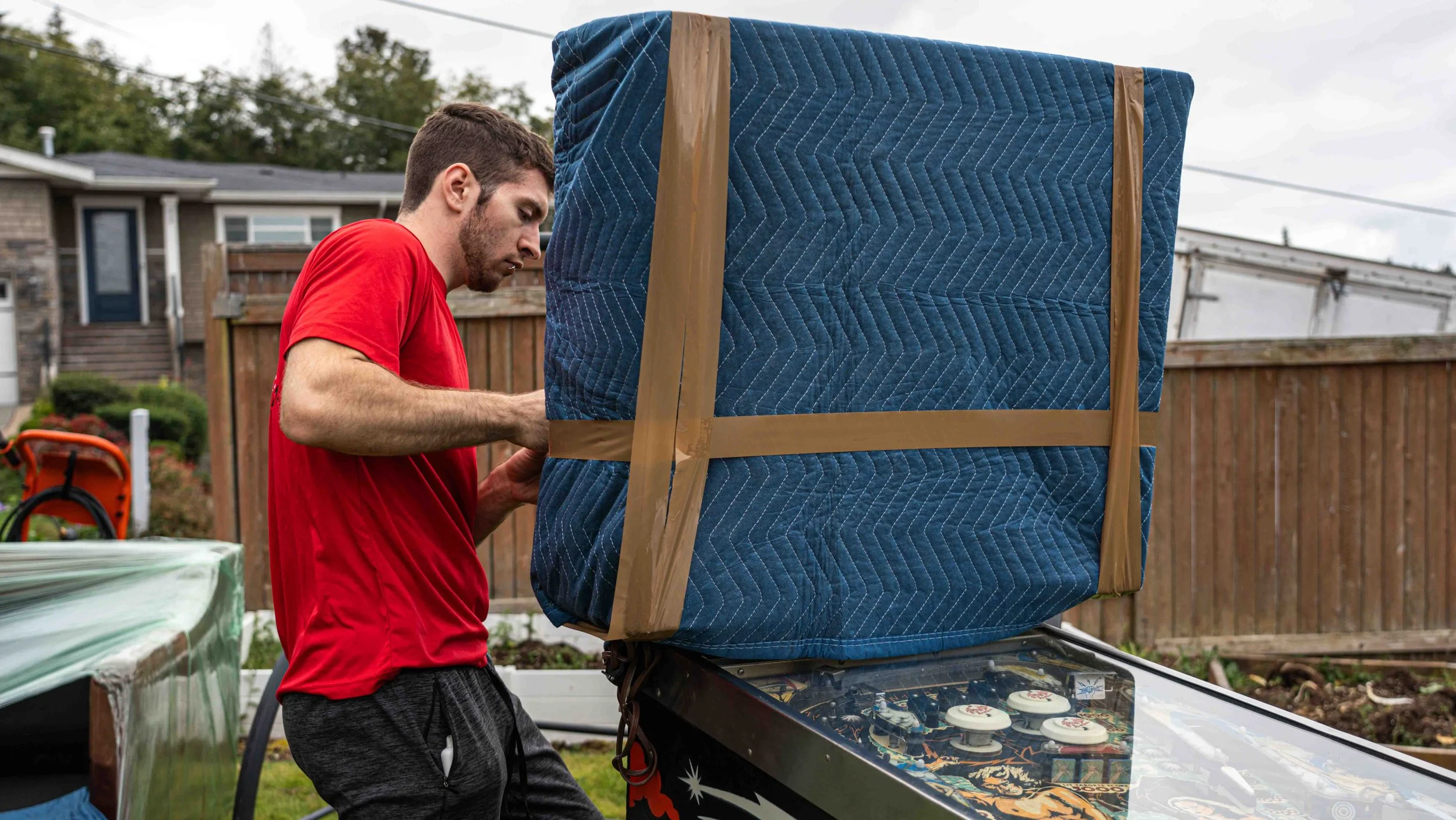 A young man in a red shirt and dark shorts is setting up a large package wrapped in blue quilted fabric on top of a car. In the background, there is a wooden fence, a house, and trees.