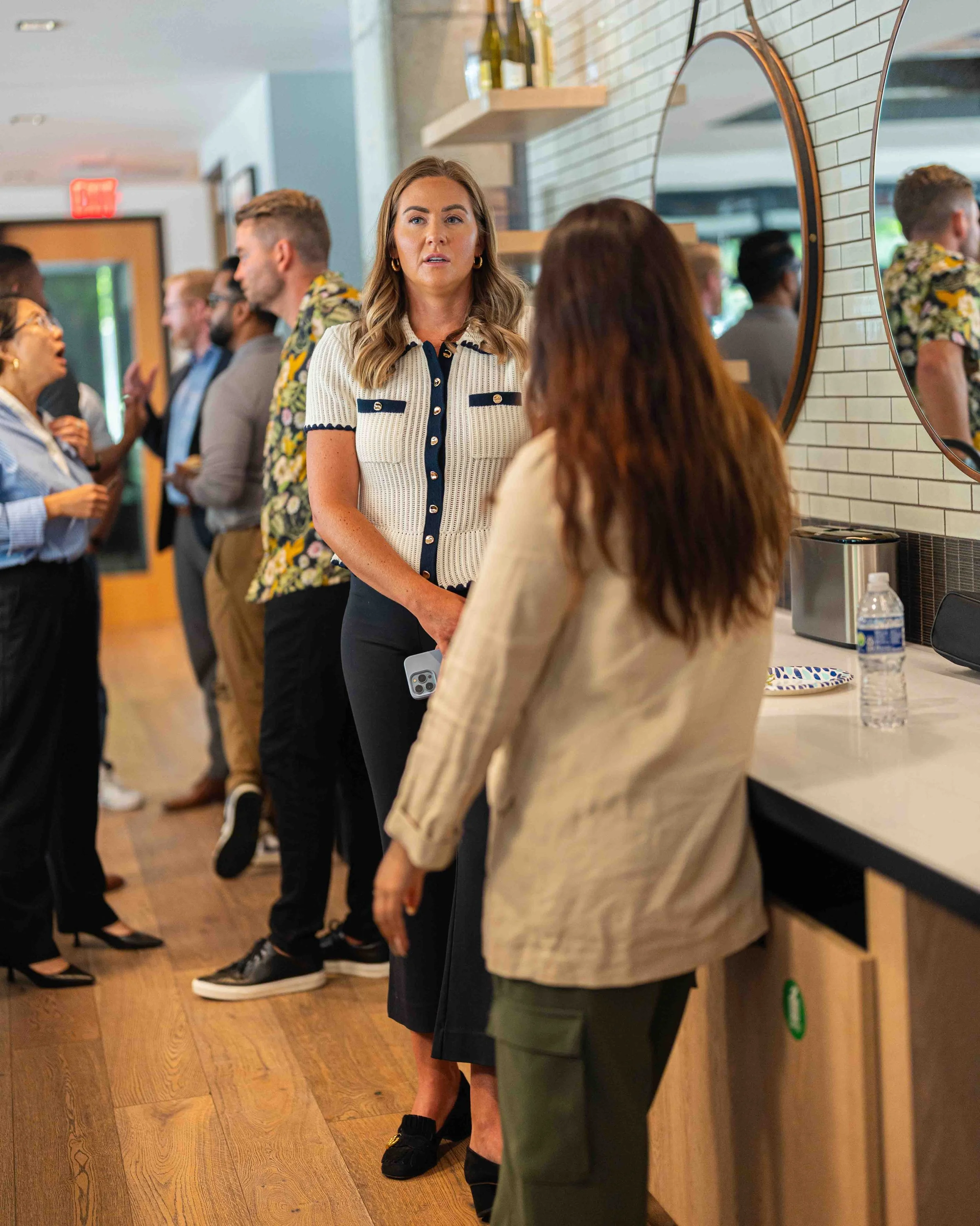 Several people engaged in conversation in a modern indoor setting with light-colored brick walls and wooden furniture. A woman in a striped shirt is at the center, while others are visible in the background and on the sides. Some individuals are look