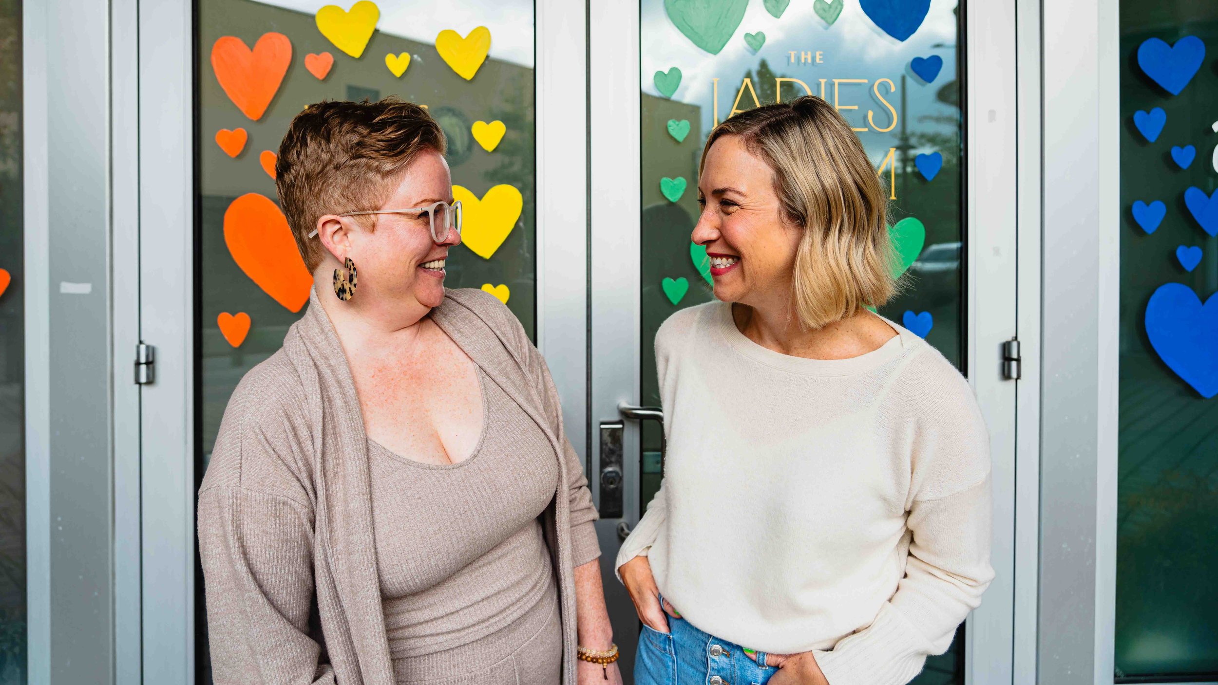 Two women smiling and talking in front of glass doors decorated with colorful paper hearts, with a sign that reads 'The Ladies' behind them.