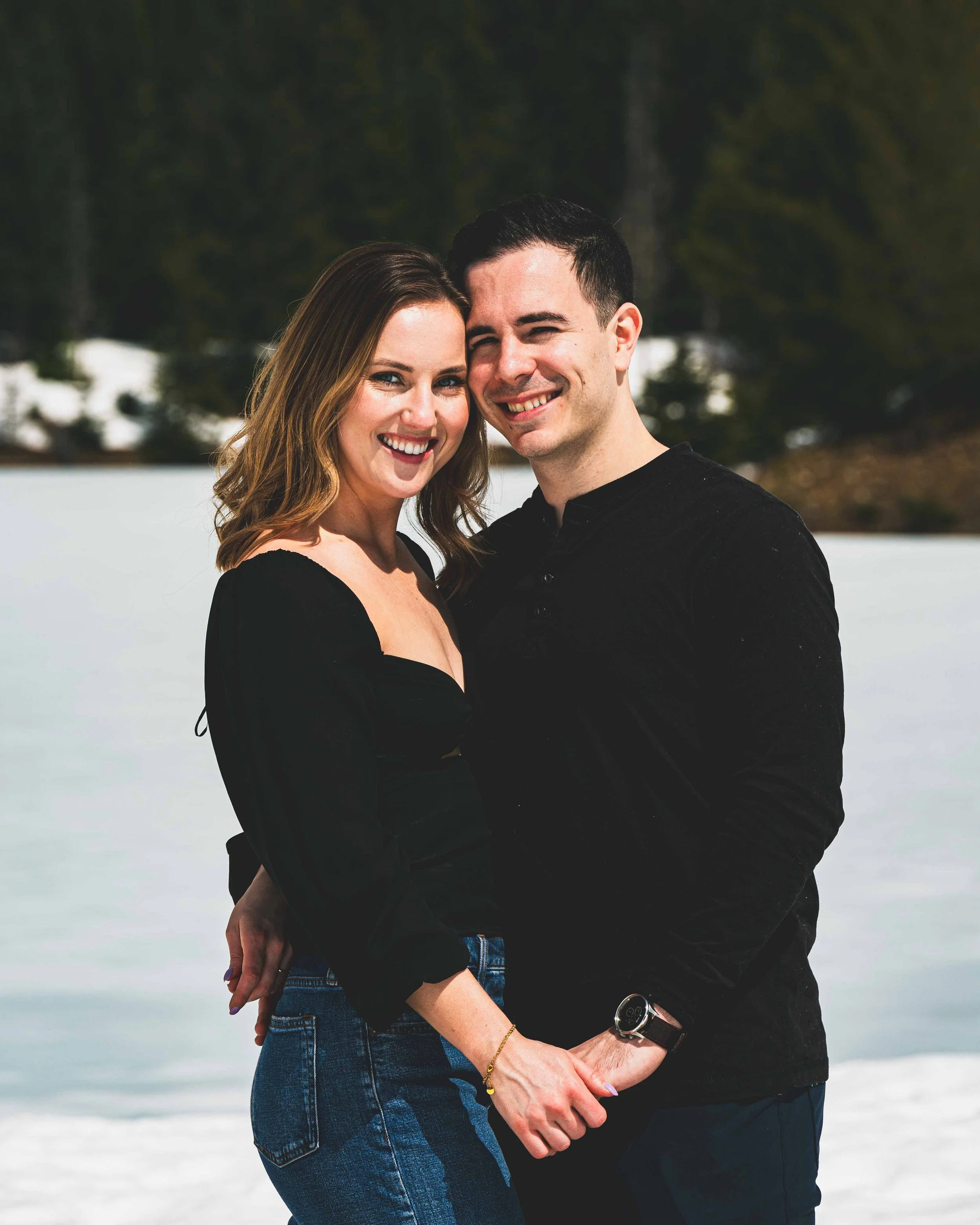 A smiling man and woman standing close together outdoors with snowy ground and trees in the background, holding hands.