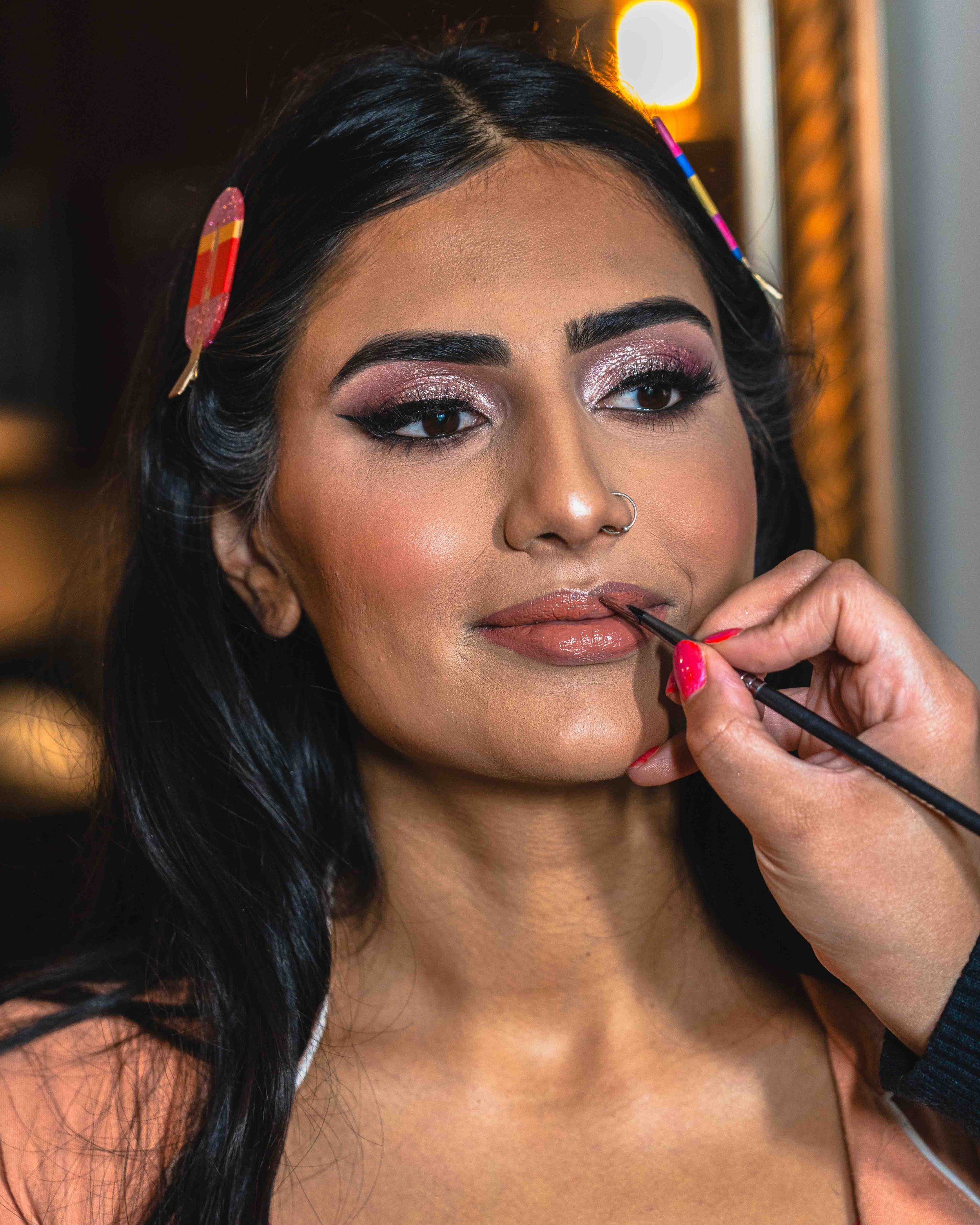 A woman with long dark hair applying lipstick with a brush. She has makeup on, including eyeshadow, eyeliner, and lipstick, and is wearing a strapless top. A makeup artist or another person is holding the lipstick brush near her lips.