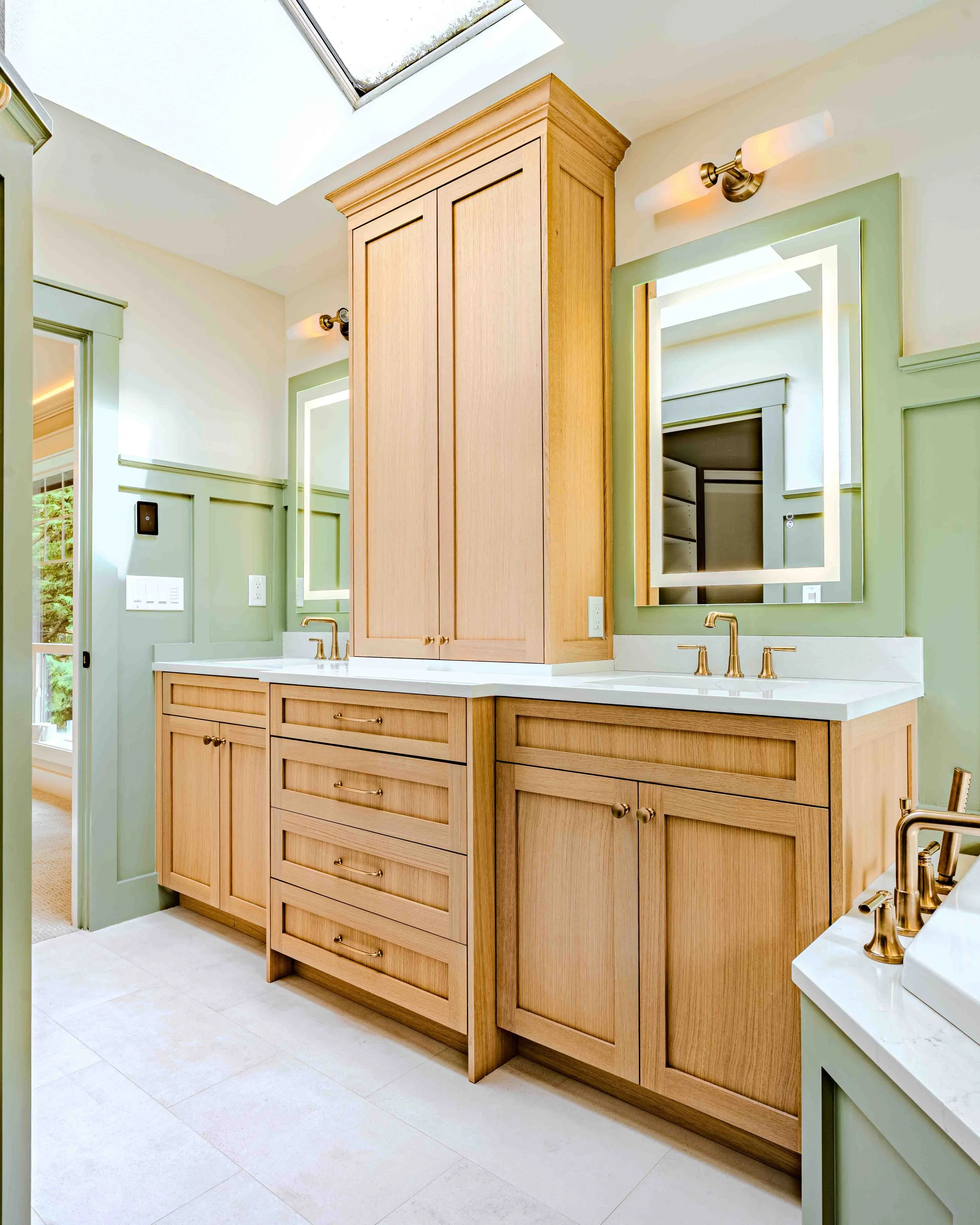A bathroom vanity with wooden cabinets, a white countertop, gold fixtures, and a large mirror. The wall is painted green, with a window and a skylight above.