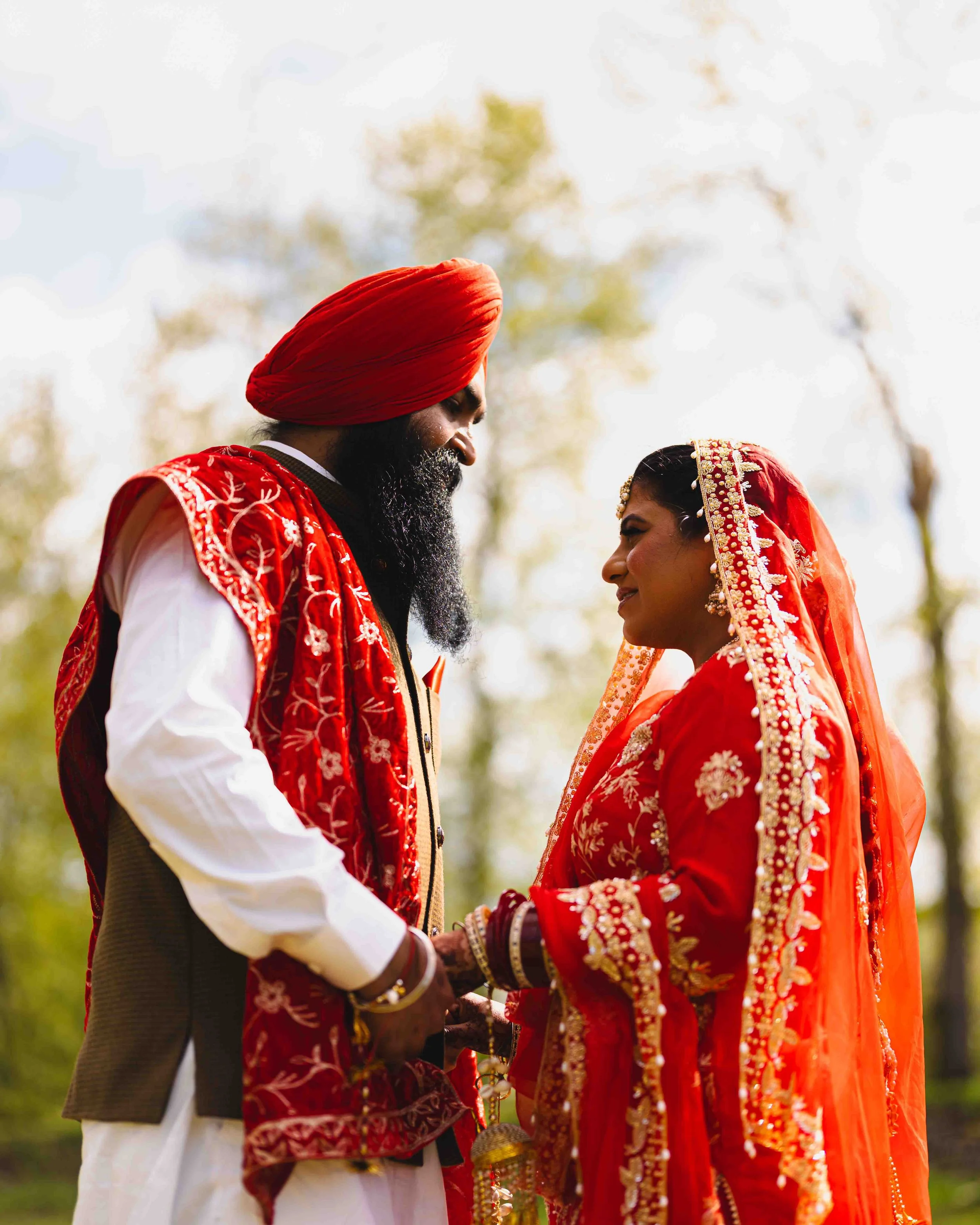 An Indian couple dressed in traditional wedding attire, standing closely and facing each other outdoors with a blurred natural background.