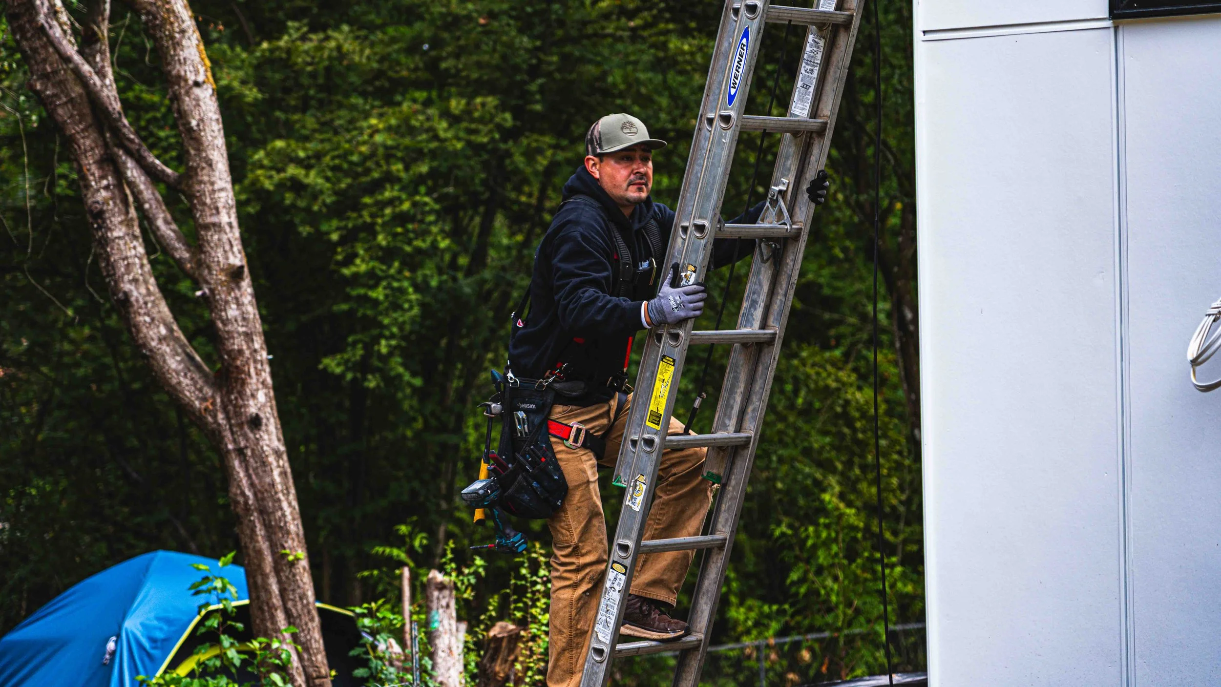 A man with tools on his belt standing on a ladder next to a white structure, outdoors with trees and a blue tent in the background.