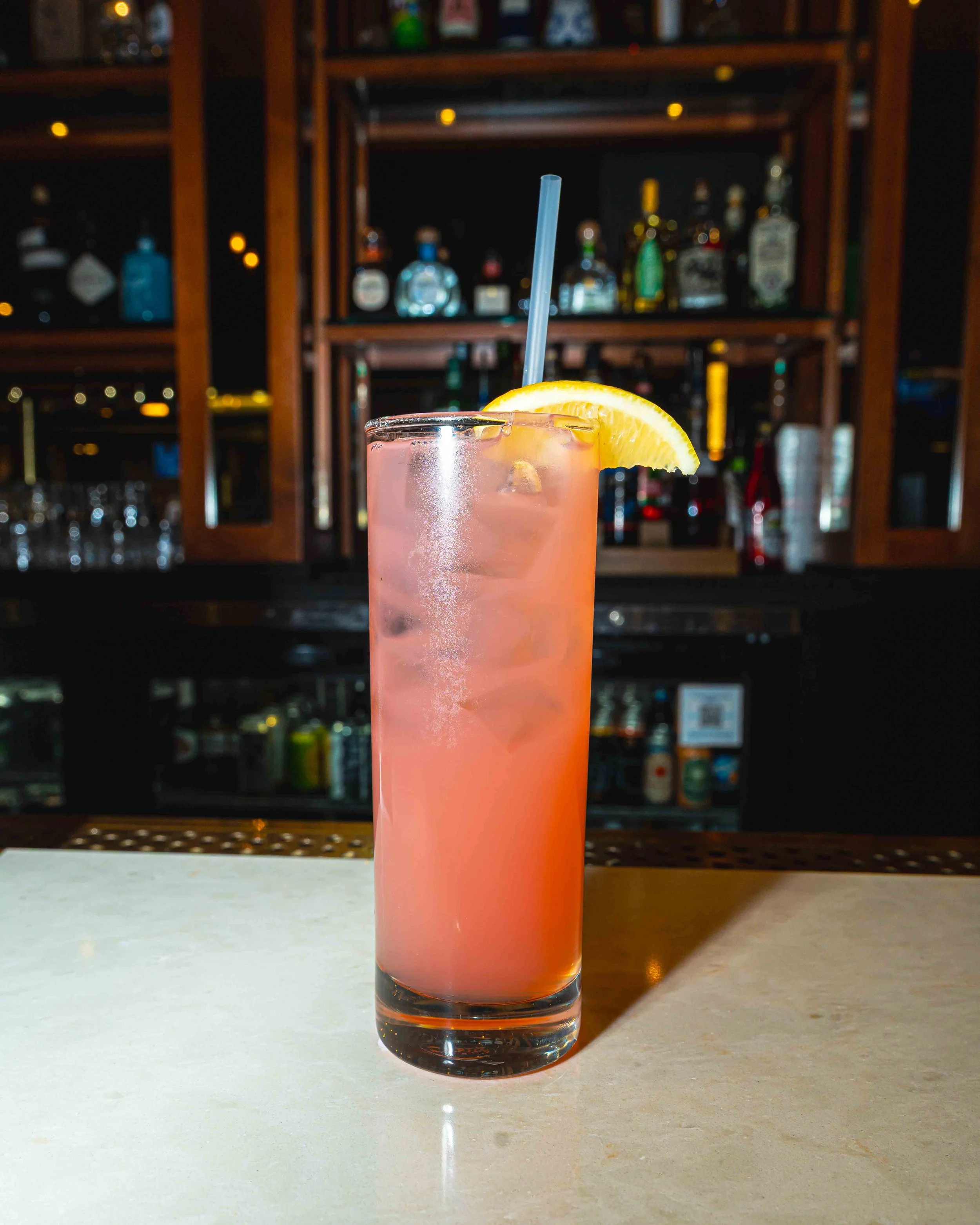 A pink cocktail with ice cubes garnished with a lemon wedge and a blue straw, on a white countertop in front of a bar with bottles on shelves.
