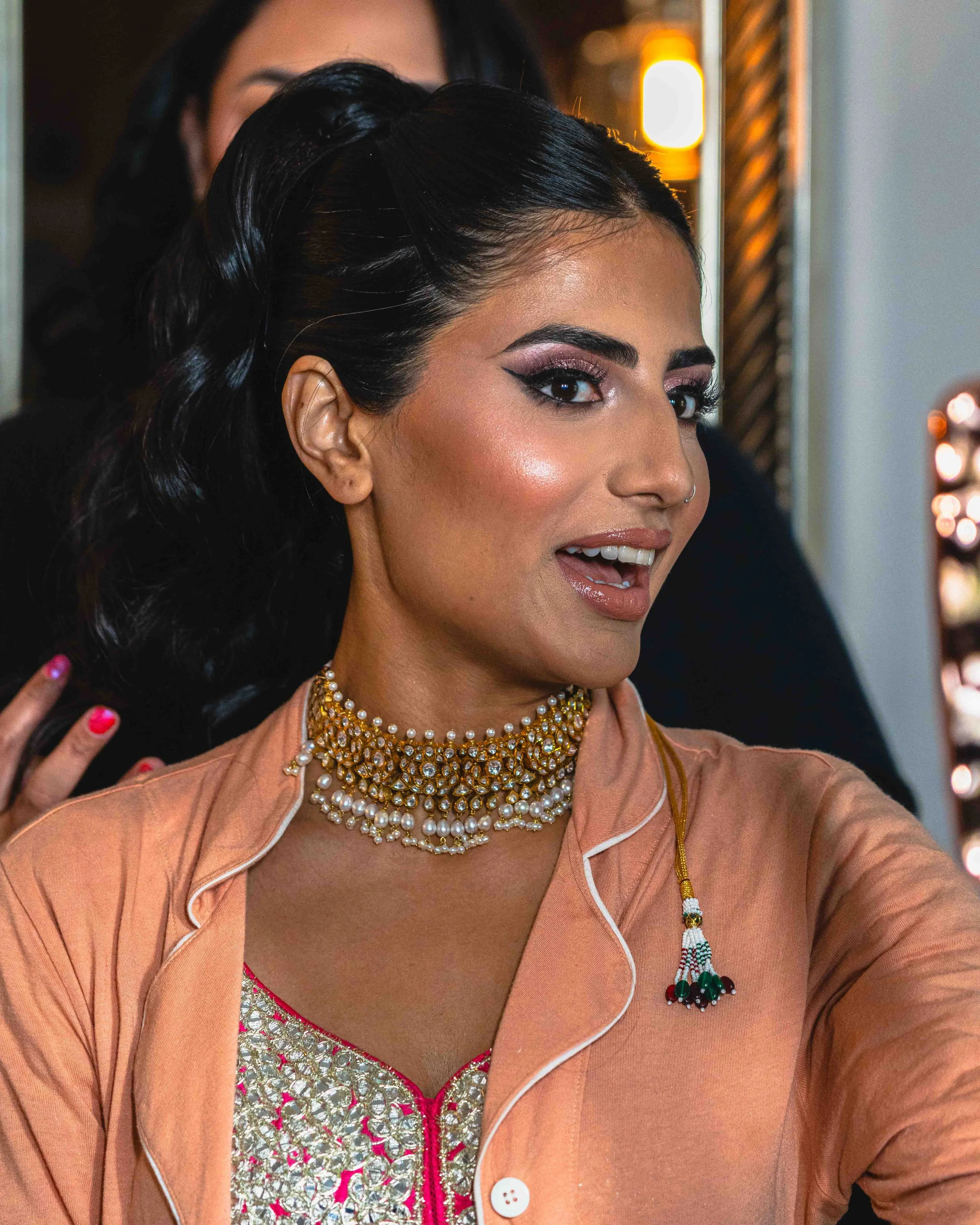Close-up of a woman with long, styled black hair, wearing makeup, a traditional Indian beaded necklace, and colorful traditional clothing with pink, gold, and silver embellishments, getting ready or posing for a photo.