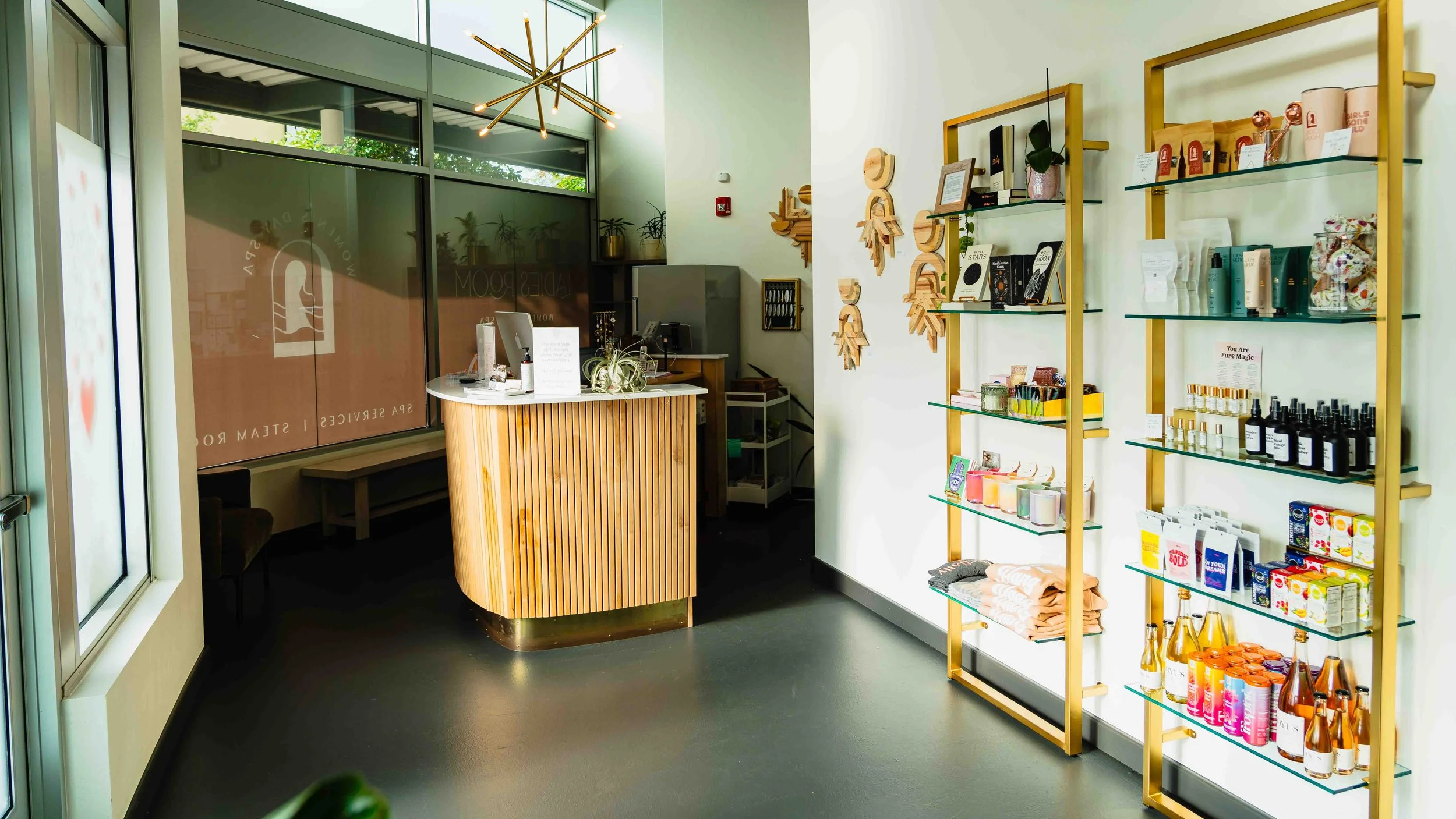 Interior of a retail store with a wooden checkout counter, display shelves with skincare and self-care products, decorative wooden wall art, and large windows allowing natural light.