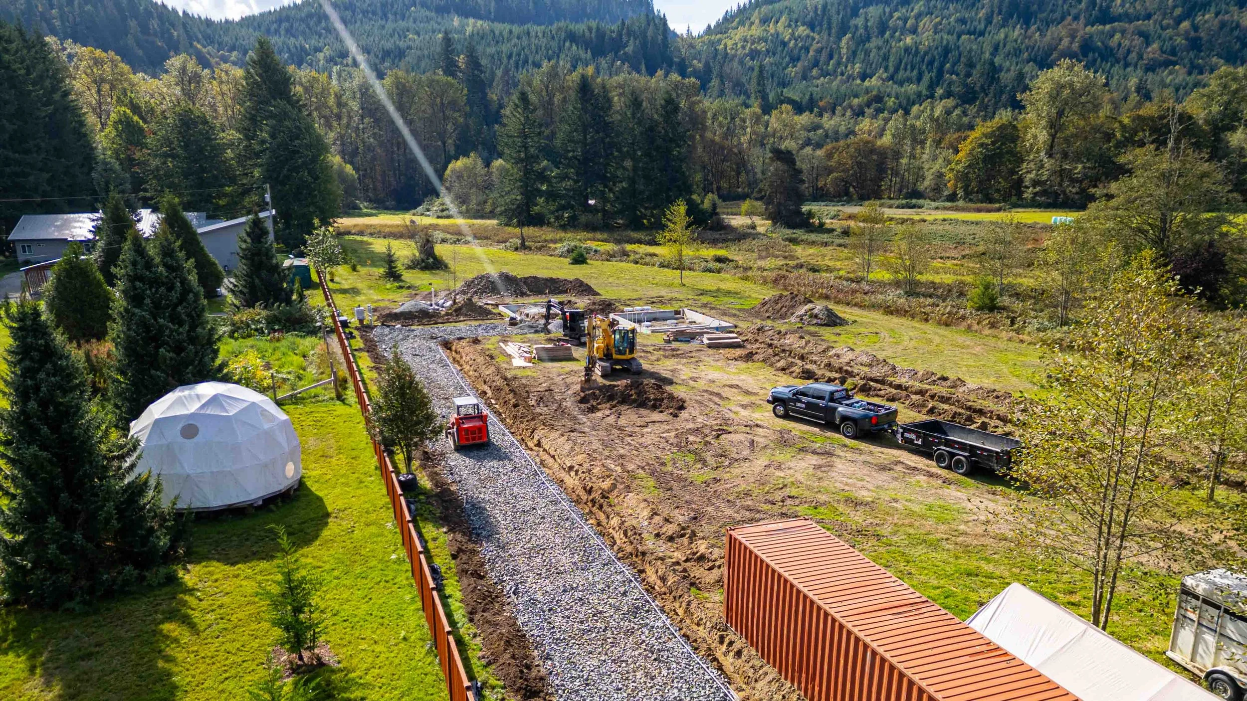 A construction site in a rural area surrounded by trees and mountains, with a small excavator, trucks, and building materials.