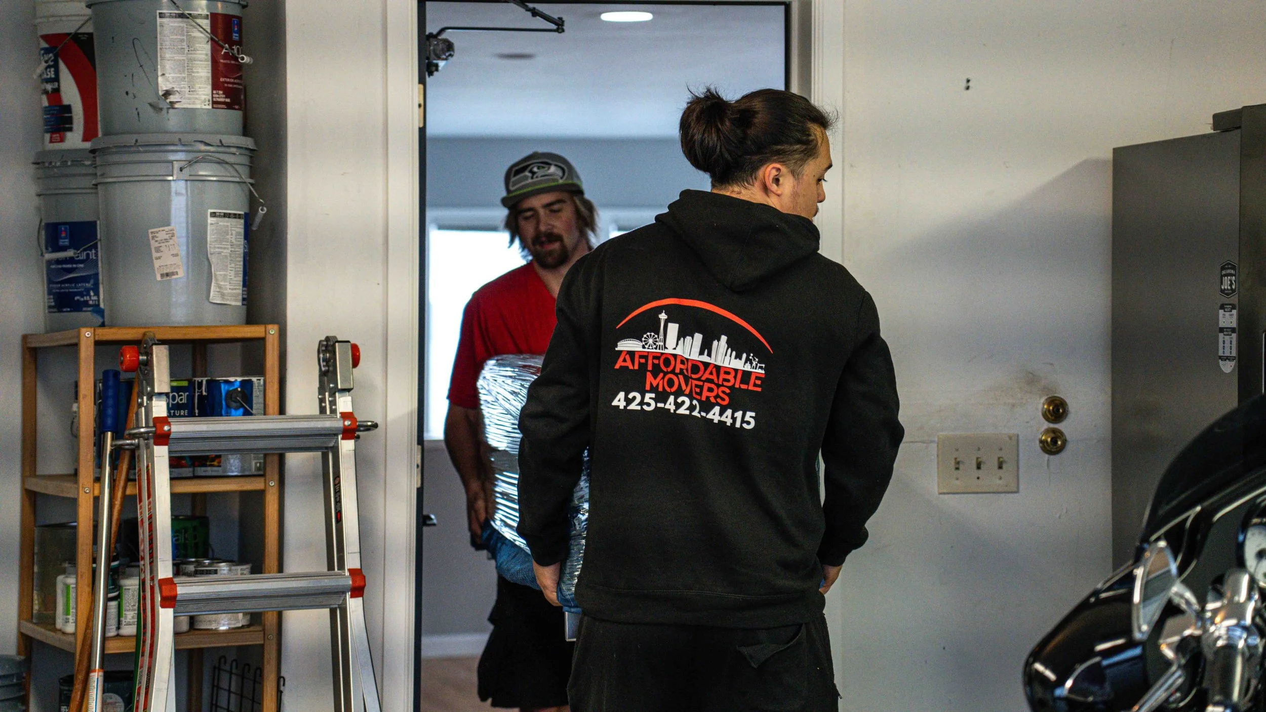 Two people are inside a garage, one of them is a woman with dark hair tied back, wearing a black hoodie with 'Affordable Movers' printed on the back. The other person, a man with long hair and a beard, is walking through the doorway carrying a wrappe