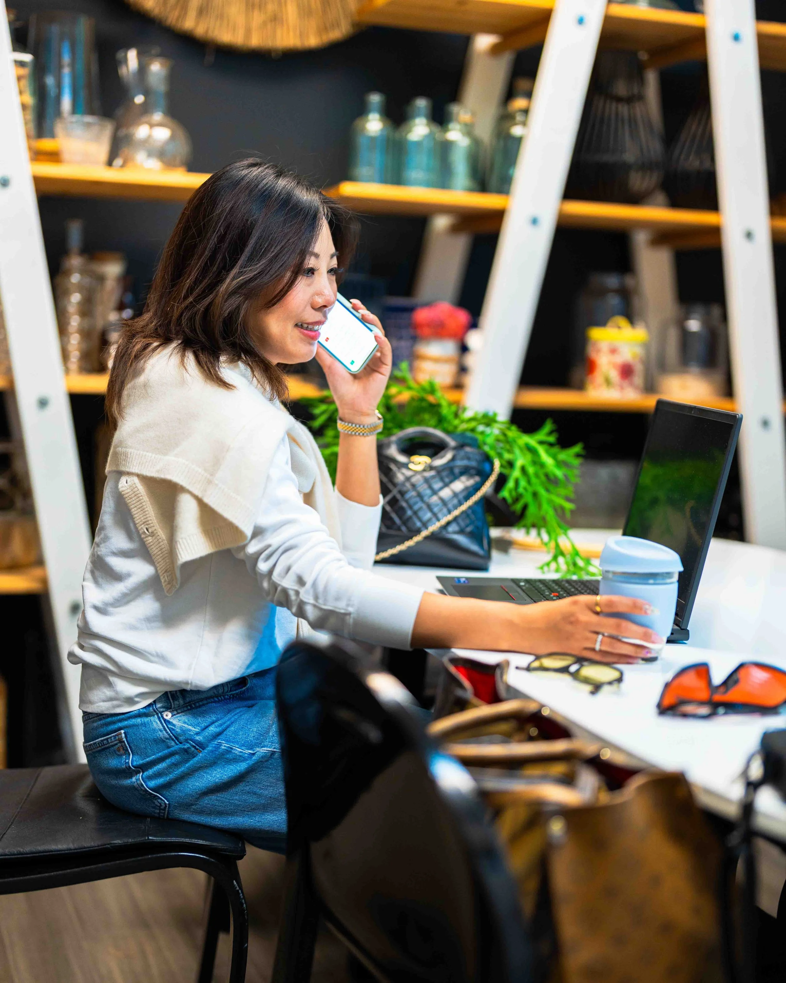 Woman kneeling on a black chair in a modern kitchen or coffee shop, talking on her smartphone while working on her laptop, with various jars and bottles on wooden shelves in the background.
