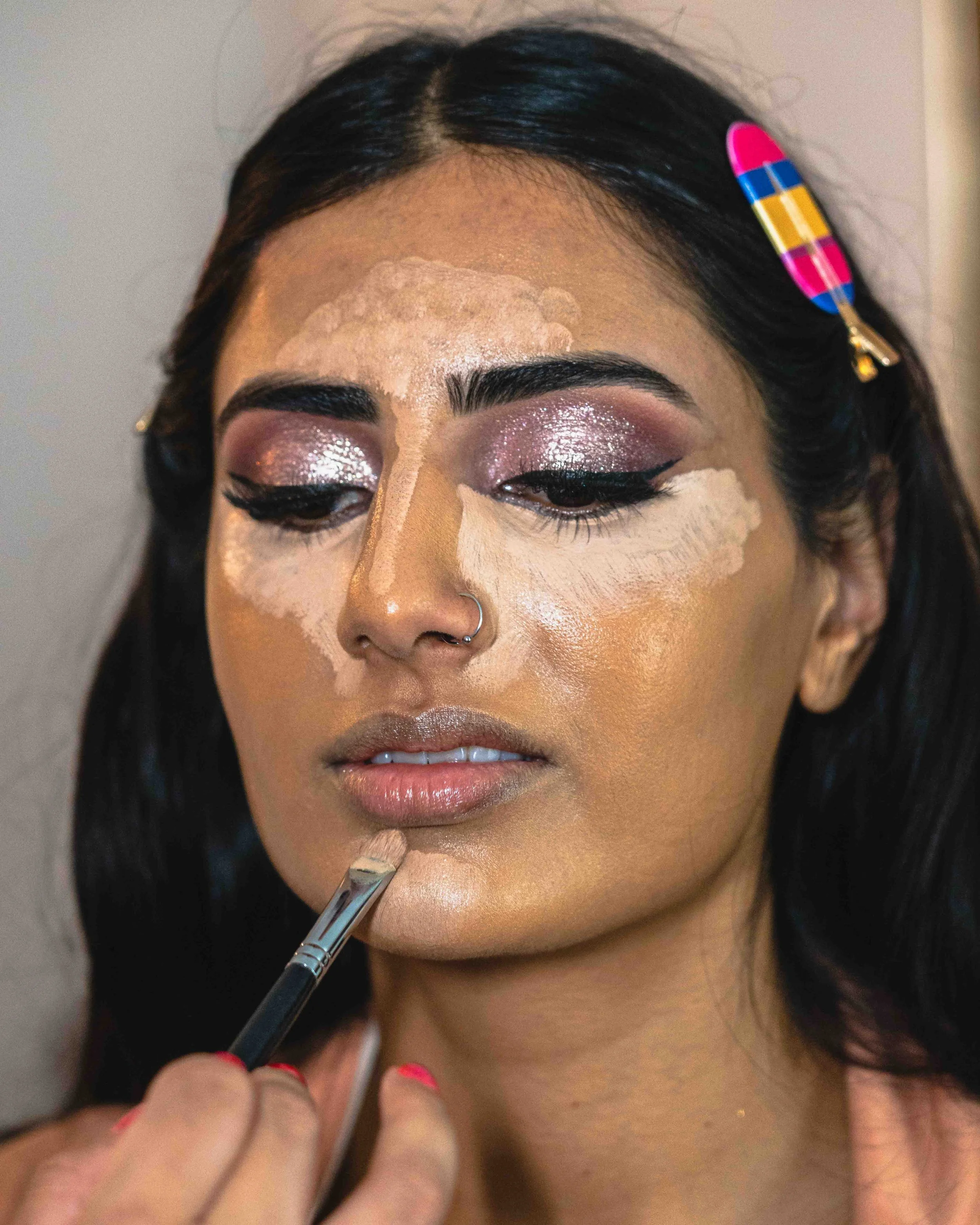 A woman with dark hair has her makeup done, with eye shadow and eyeliner. A person is applying foundation to her face with a brush, using light-colored makeup and concealer around the eyes and forehead.