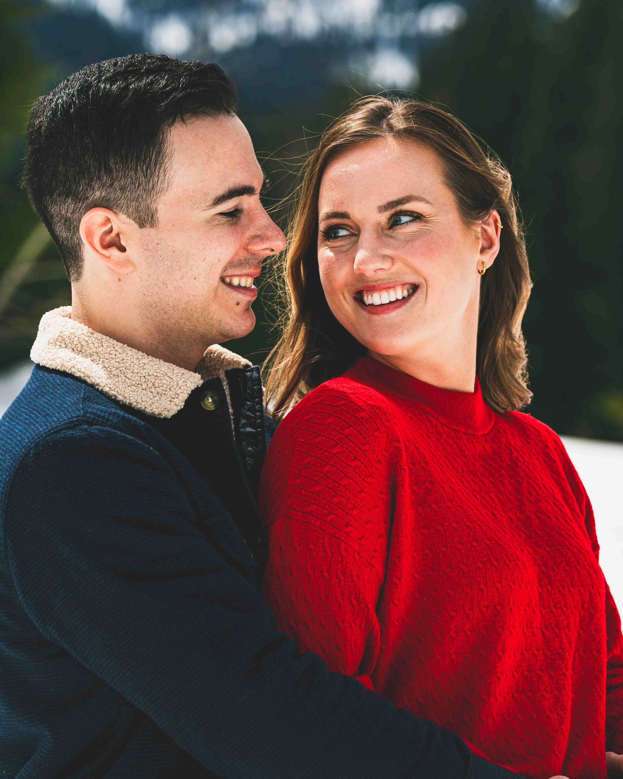 A close-up of a smiling man and woman looking at each other outdoors. The man has dark hair and is wearing a navy jacket with a sherpa collar. The woman has light brown hair and is wearing a red textured sweater. They are standing close together with