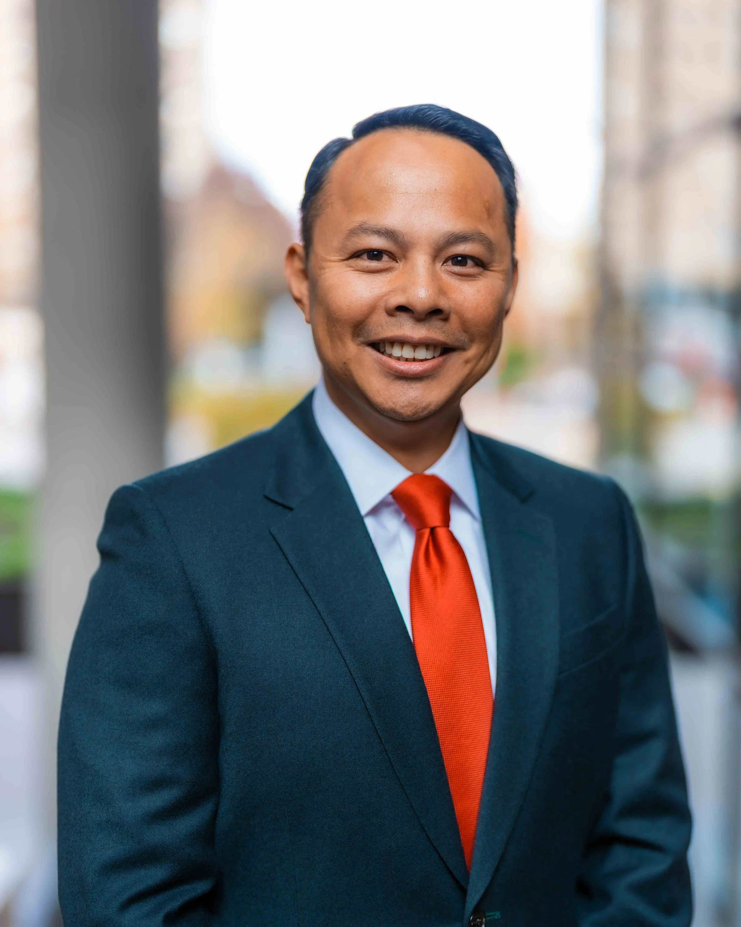 Close-up of a smiling man in a business suit with a red tie, standing outdoors with blurred cityscape background.