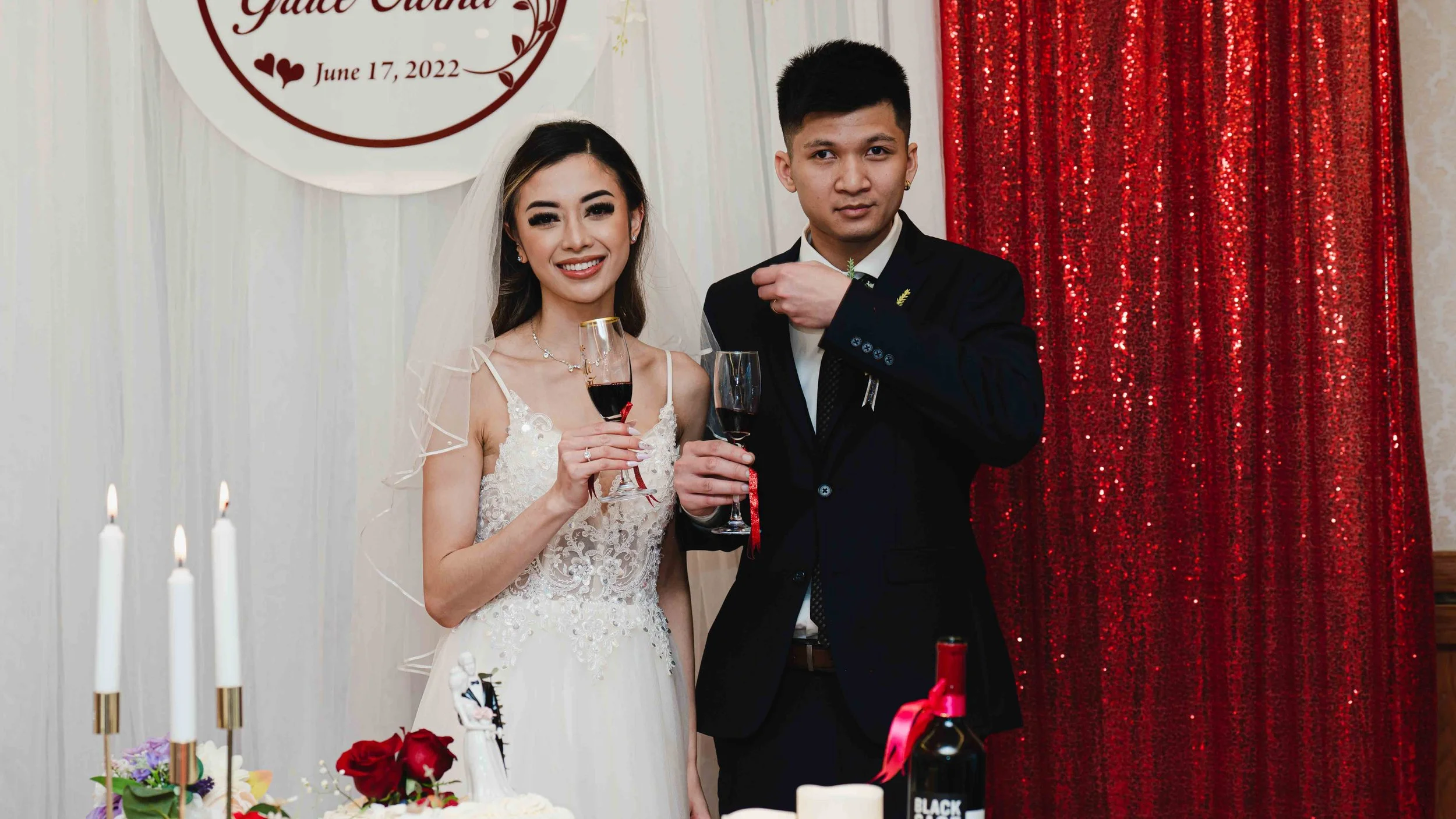 Bride and groom at their wedding reception, holding glasses of red wine and smiling. The bride is in a lace wedding dress and veil, the groom in a black suit. There are candles, flowers, and a red sequin backdrop with a white circle sign that reads "