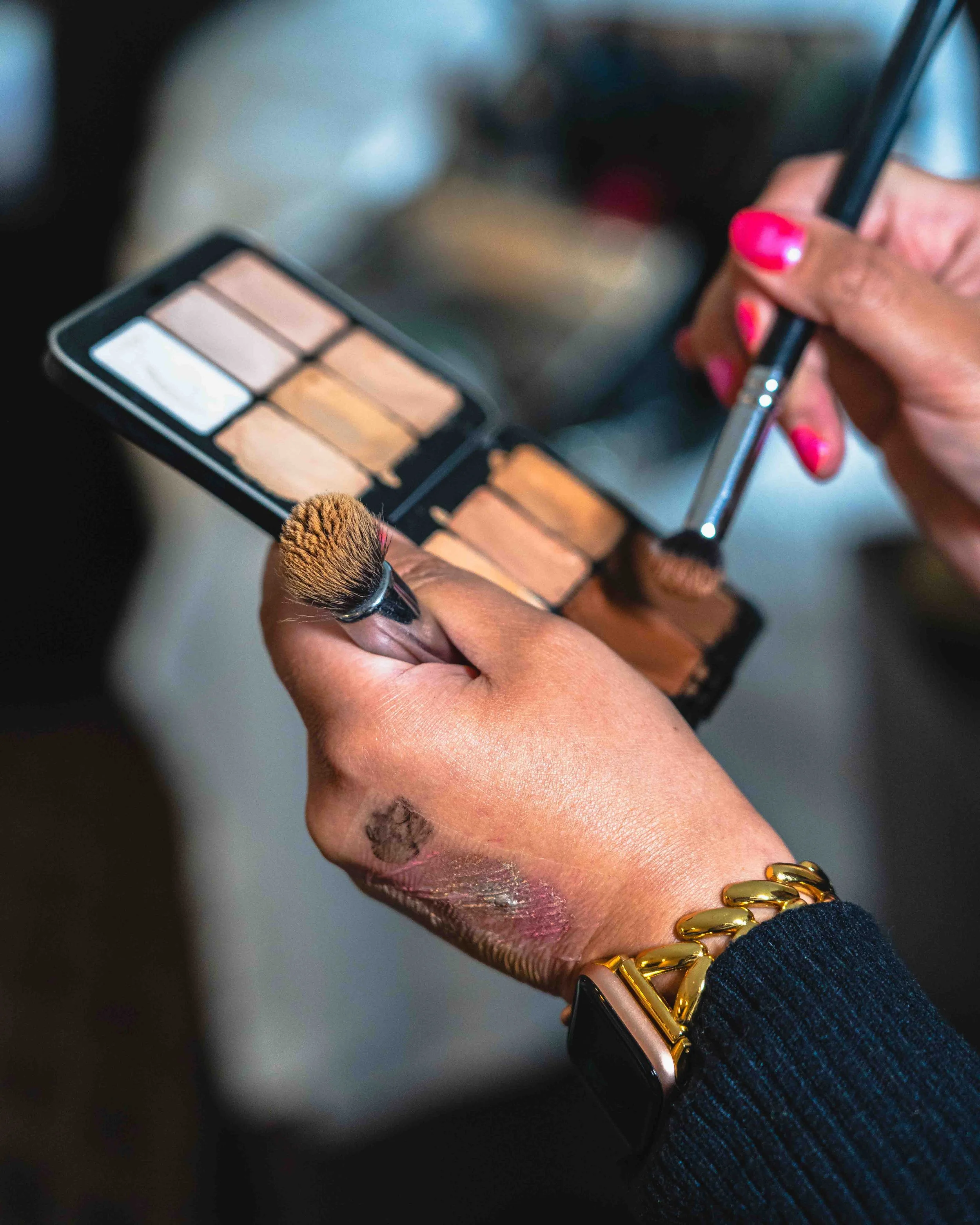 Close-up of a woman's hand holding a makeup brush and a palette of foundation shades, with pink nail polish and wearing a black bracelet with gold links.