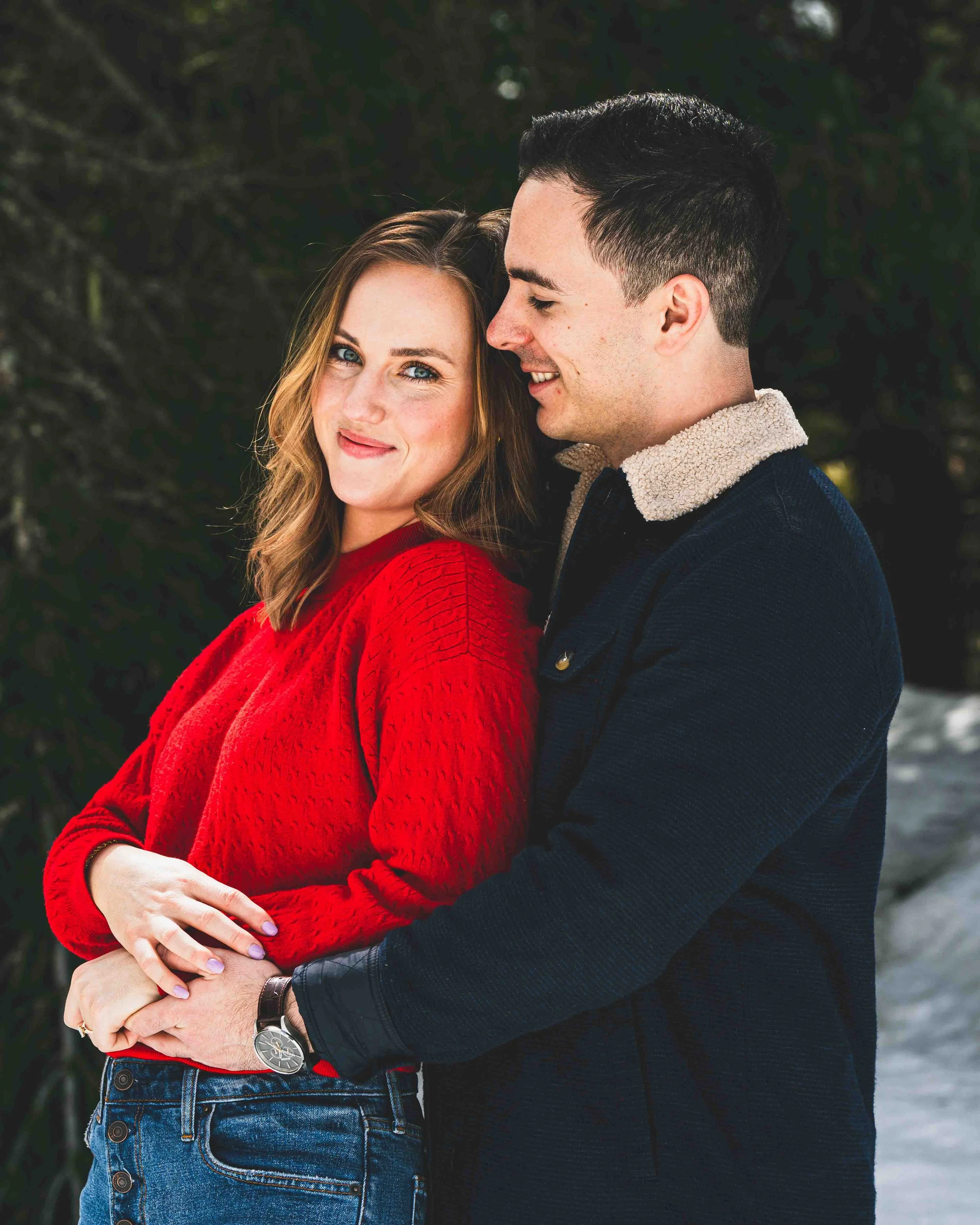 A couple embracing outdoors, with the woman wearing a red sweater and the man wearing a dark jacket with a beige collar. They are close together, smiling softly, with the man's arm around the woman's waist.