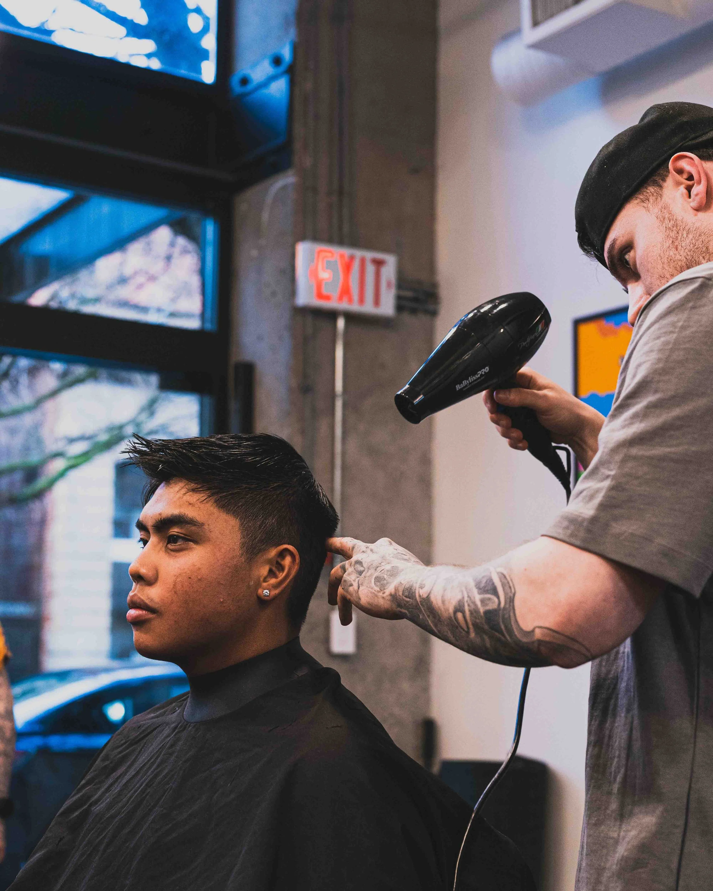 A man with short dark hair and earrings is sitting in a salon while a hairstylist blow-dries his hair. The stylist has tattoos on their forearm and is using a black hairdryer.