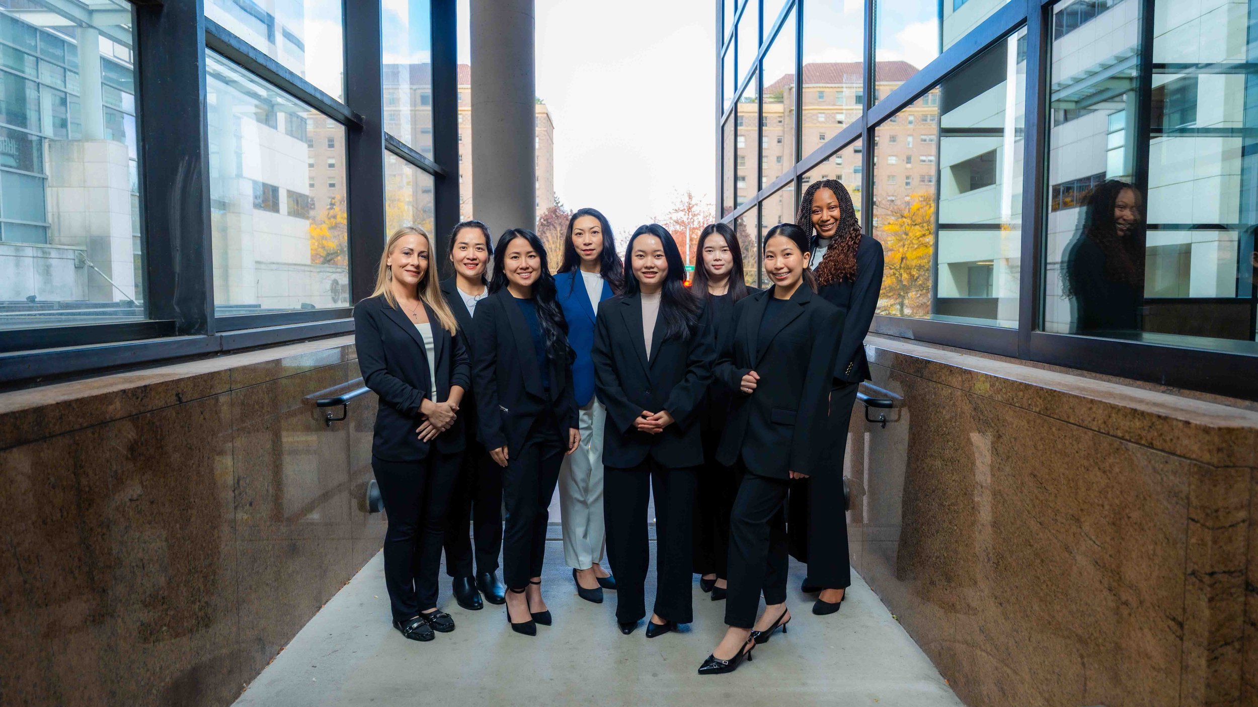 Group of nine women in business attire standing together outdoors near a glass building with cityscape in the background.
