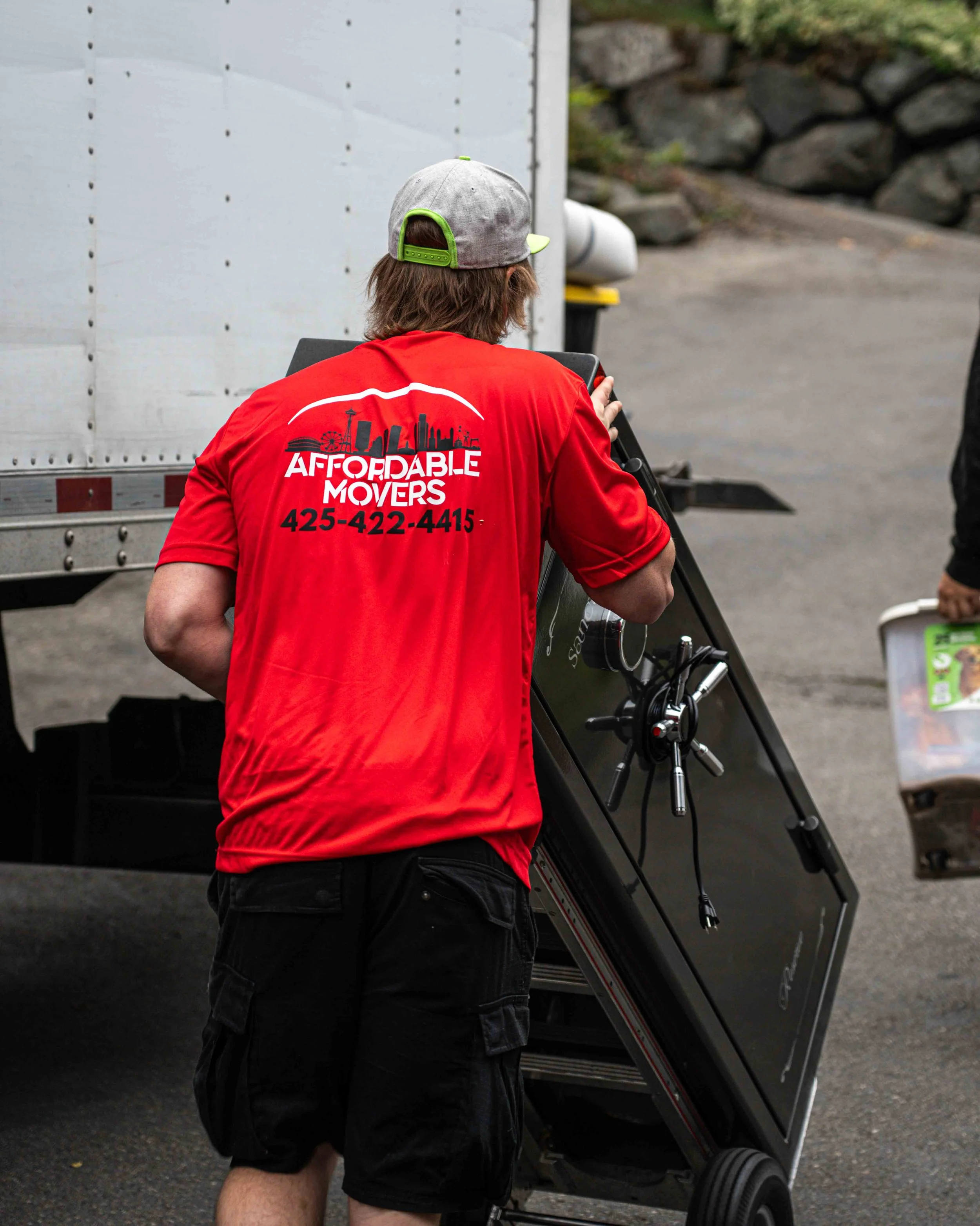 Person with brown hair wearing a gray baseball cap and a red shirt with 'Affordable Movers' and a phone number, loading or unloading a black moving dolly or cart near a white truck.