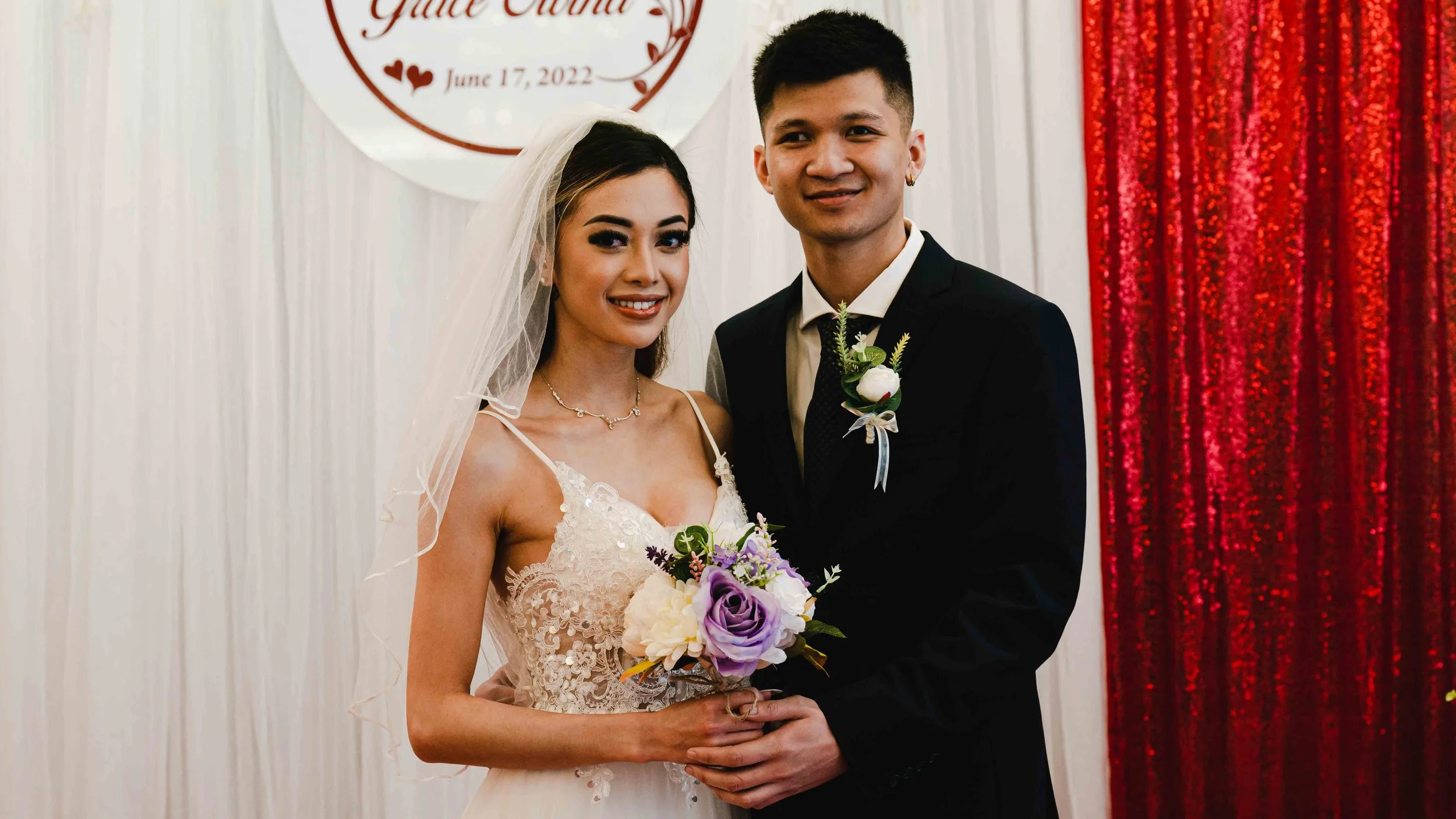 A bride and groom pose together at their wedding, with the bride holding a bouquet of flowers and the background decorated with a sign that has the date June 17, 2022, and the couple's wedding name.