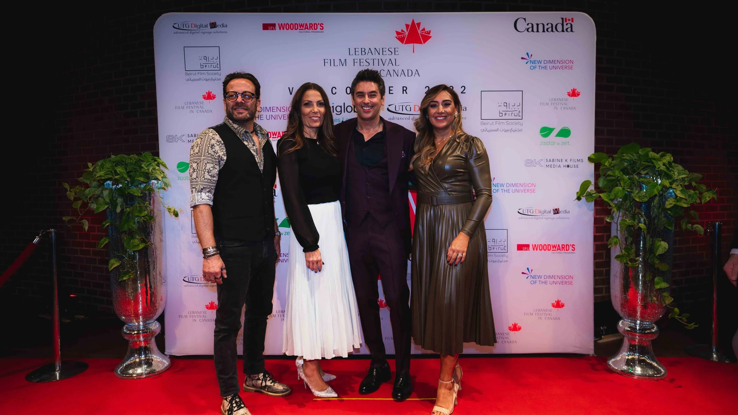 Four people smiling and standing on a red carpet at the Lebanese Film Festival in Canada, with a white backdrop featuring logos and text behind them. Two women and two men, dressed in formal attire.