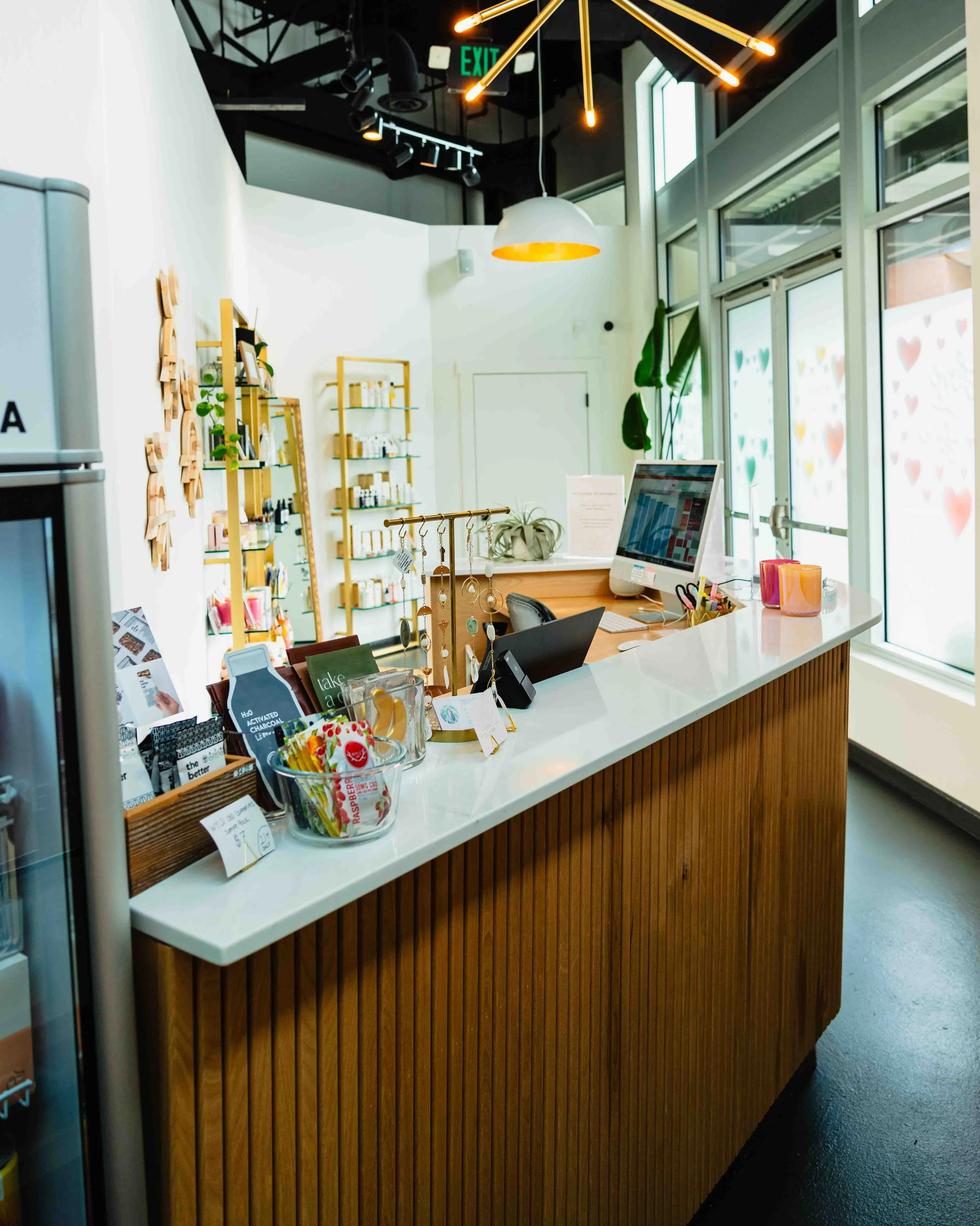 Interior of a modern retail store with a white checkout counter made of wood with vertical slats, a computer monitor, small racks of jewelry, and various retail items. Behind the counter are shelves with products, large windows with colorful heart de