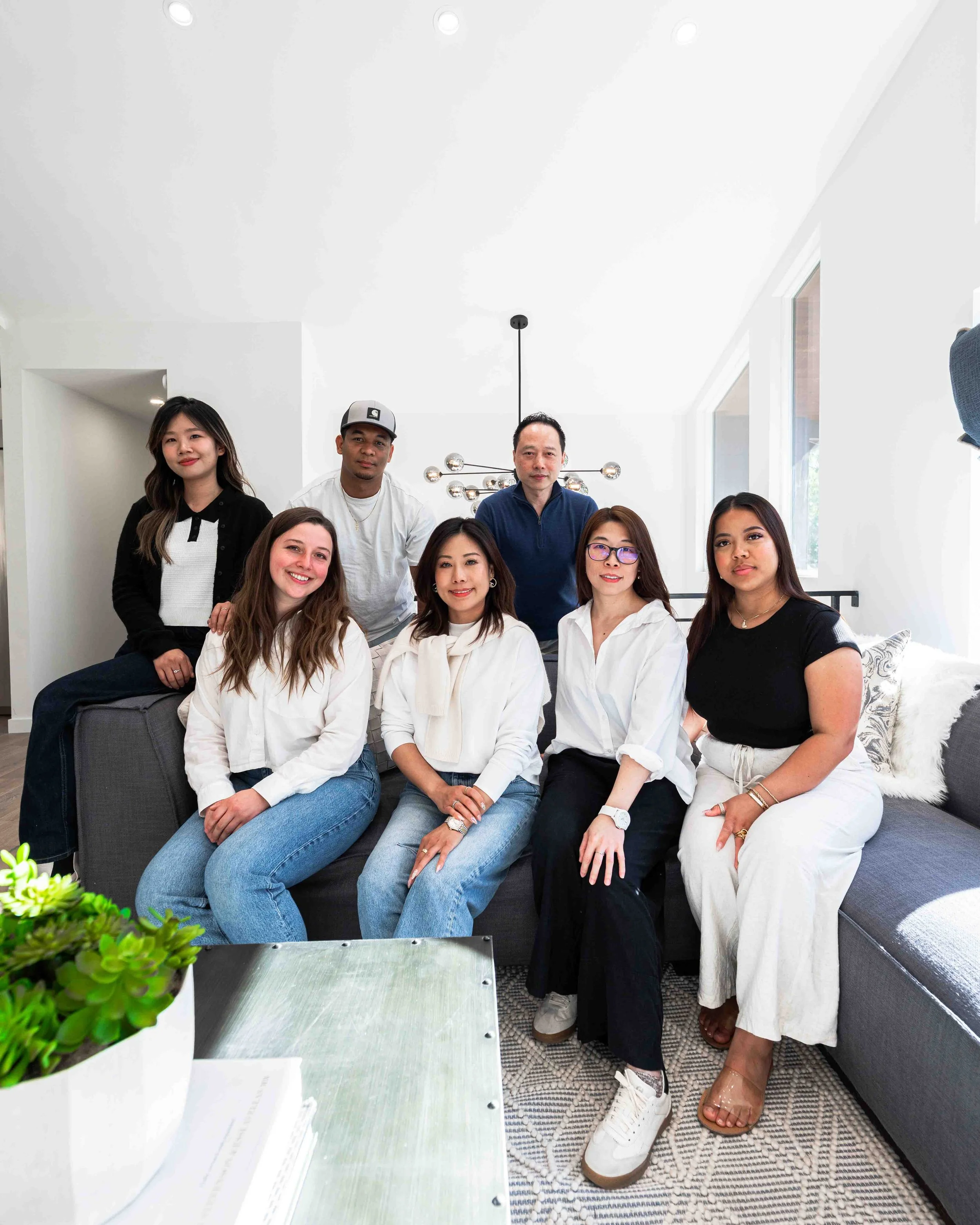 A diverse group of nine people, six women and three men, gathered in a bright, modern living room. They are sitting and standing around each other, smiling and posing for the camera. The room has large windows, a stylish chandelier, and a patterned r