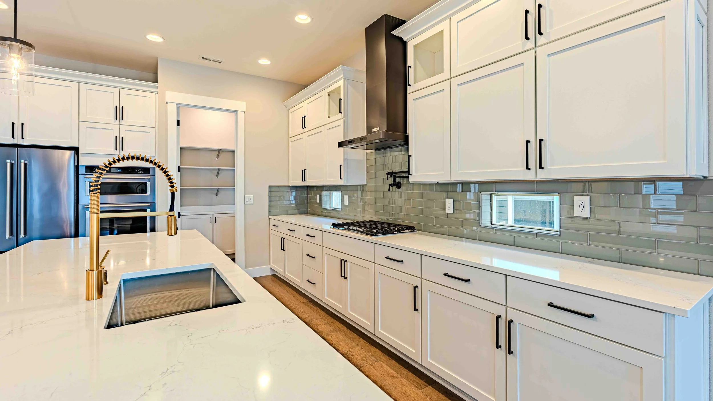 Modern kitchen with white cabinets, a marble countertop, a stainless steel refrigerator, a built-in oven, and a gas stove with a black range hood. There is a small window on the backsplash and a gold faucet with a matching sink on the island.