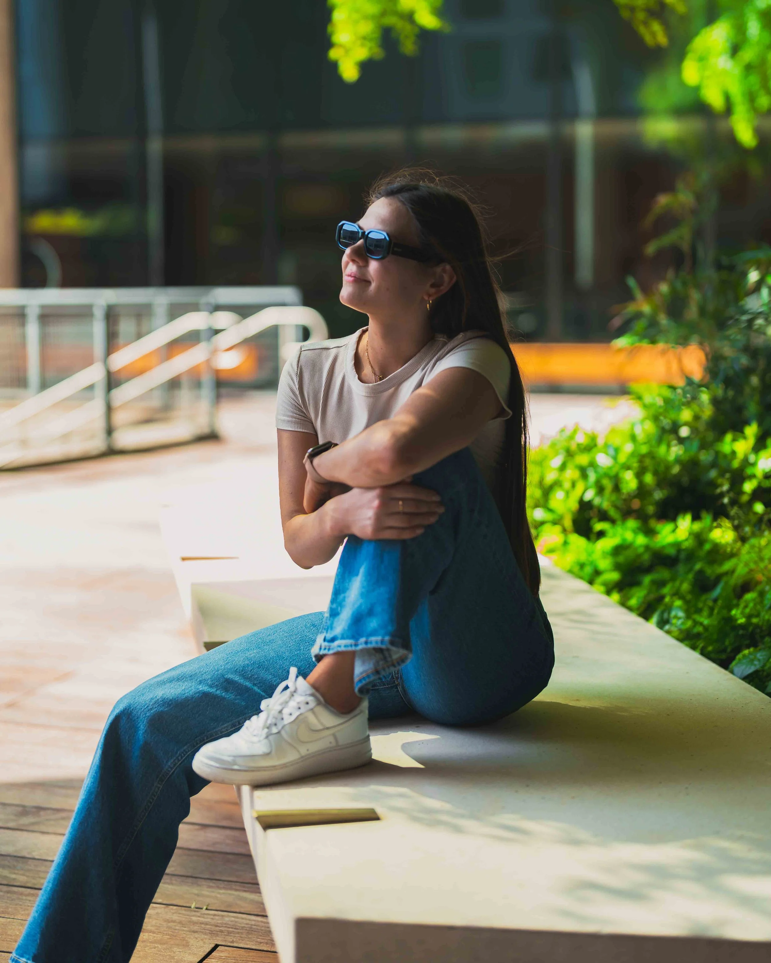 A woman is sitting on a bench outdoors, wearing dark sunglasses, a light-colored t-shirt, and blue jeans. She appears relaxed with her arms crossed and is smiling slightly. The background includes some greenery and a modern building.