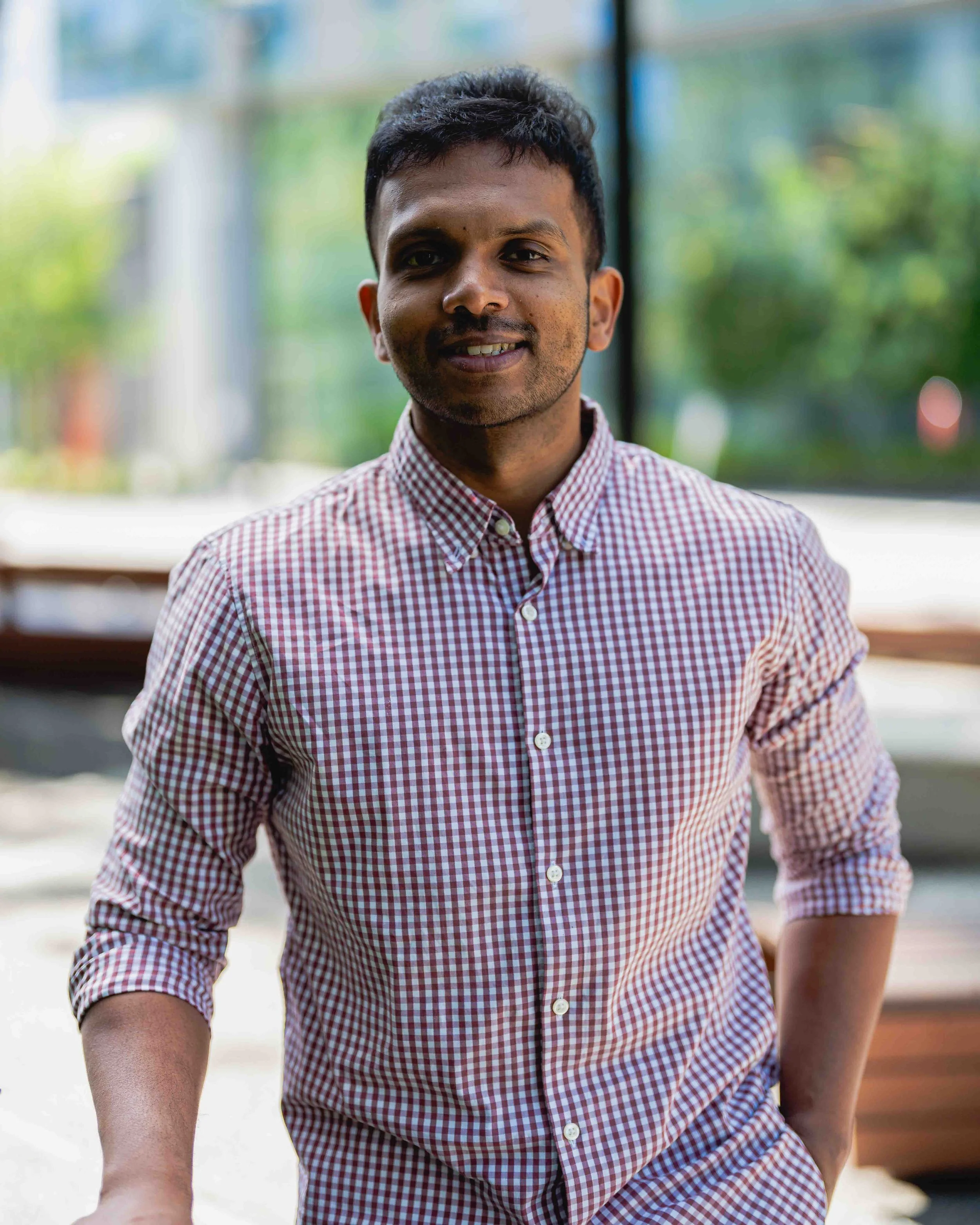 A young man with dark hair and a light beard, wearing a red and white checkered button-up shirt, smiling softly at the camera, standing outdoors with blurry green foliage in the background.