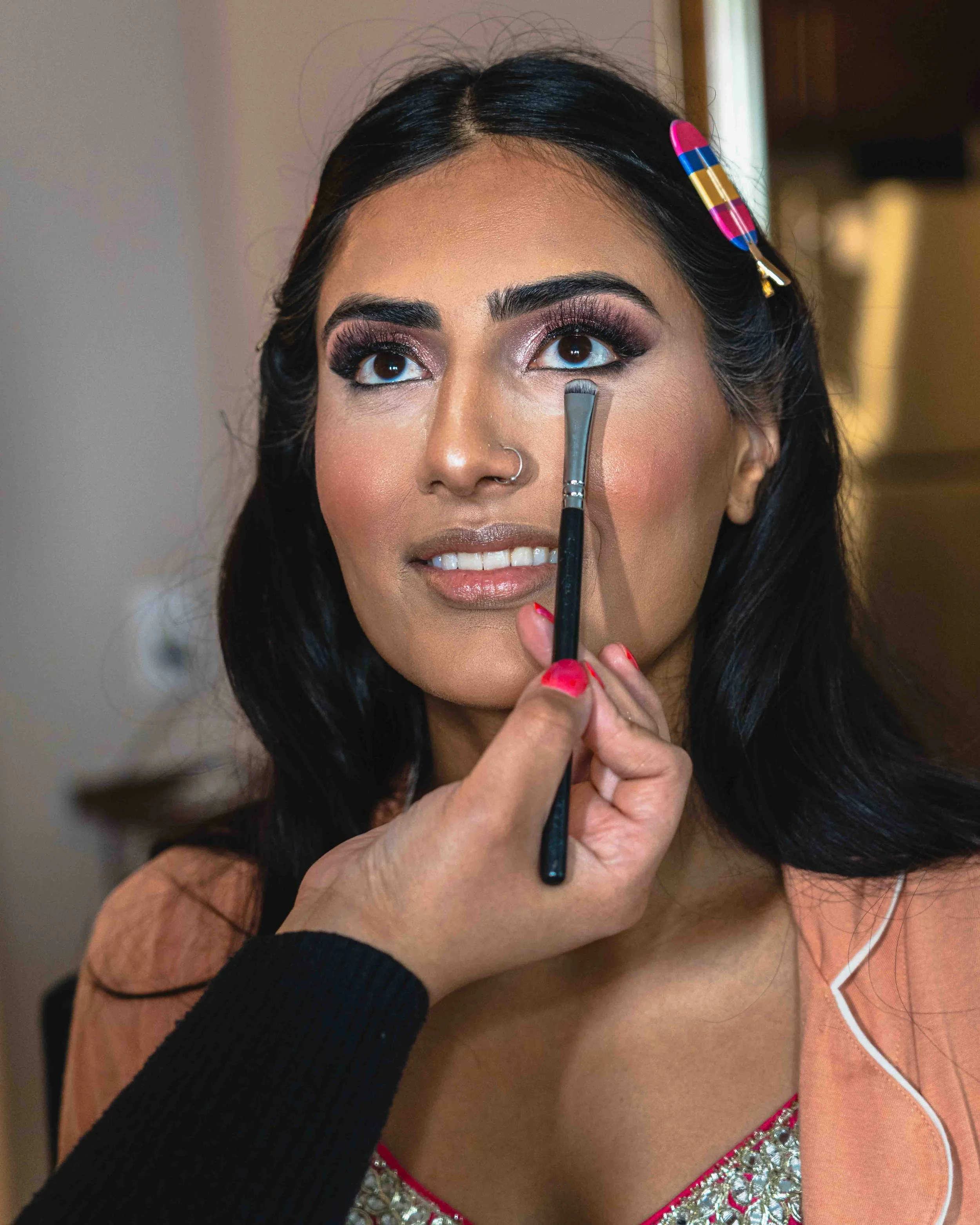 A woman with dark hair being assisted with makeup, specifically eyeshadow, by a person wearing a black long-sleeve shirt. The woman is wearing a peach-colored top with white piping and a beaded neckline.